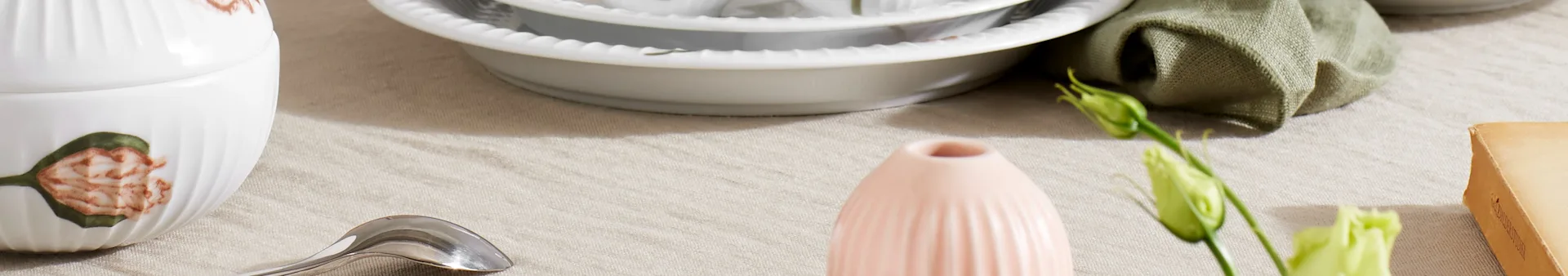 A table setting with a white ridged lidded dish, stacked white plates, a pink vase, a green flower bud, a silver spoon, and a folded green napkin on a light tablecloth.