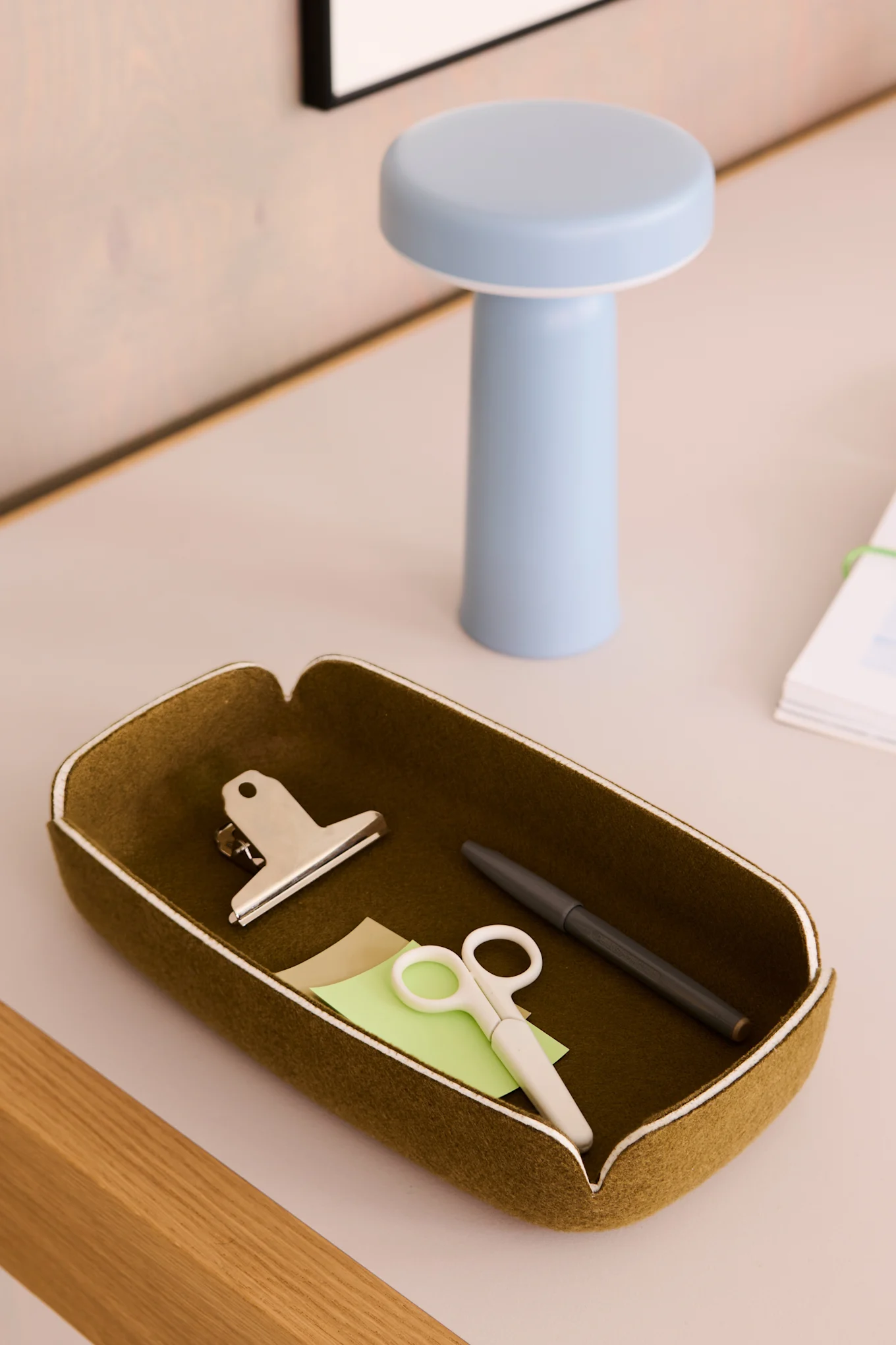 An olive green felt desk organizer tray containing a silver clip, white scissors, a grey pen, and sticky notes, with a blue lamp in the background.