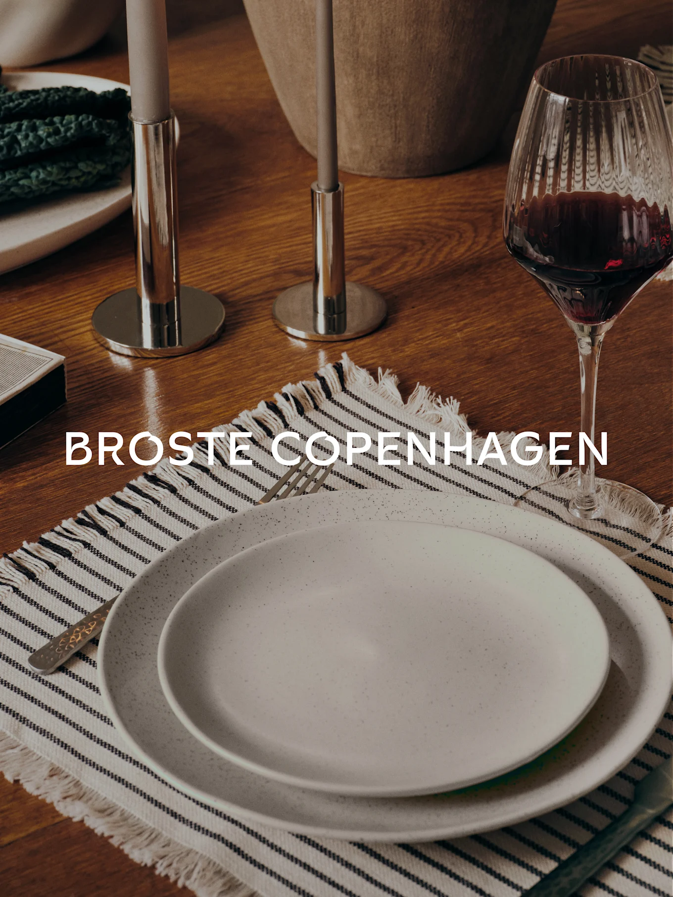 Dining table with two speckled plates on a striped placemat, fork, wine glass, two candlesticks, and 'BROSTE COPENHAGEN' text.
