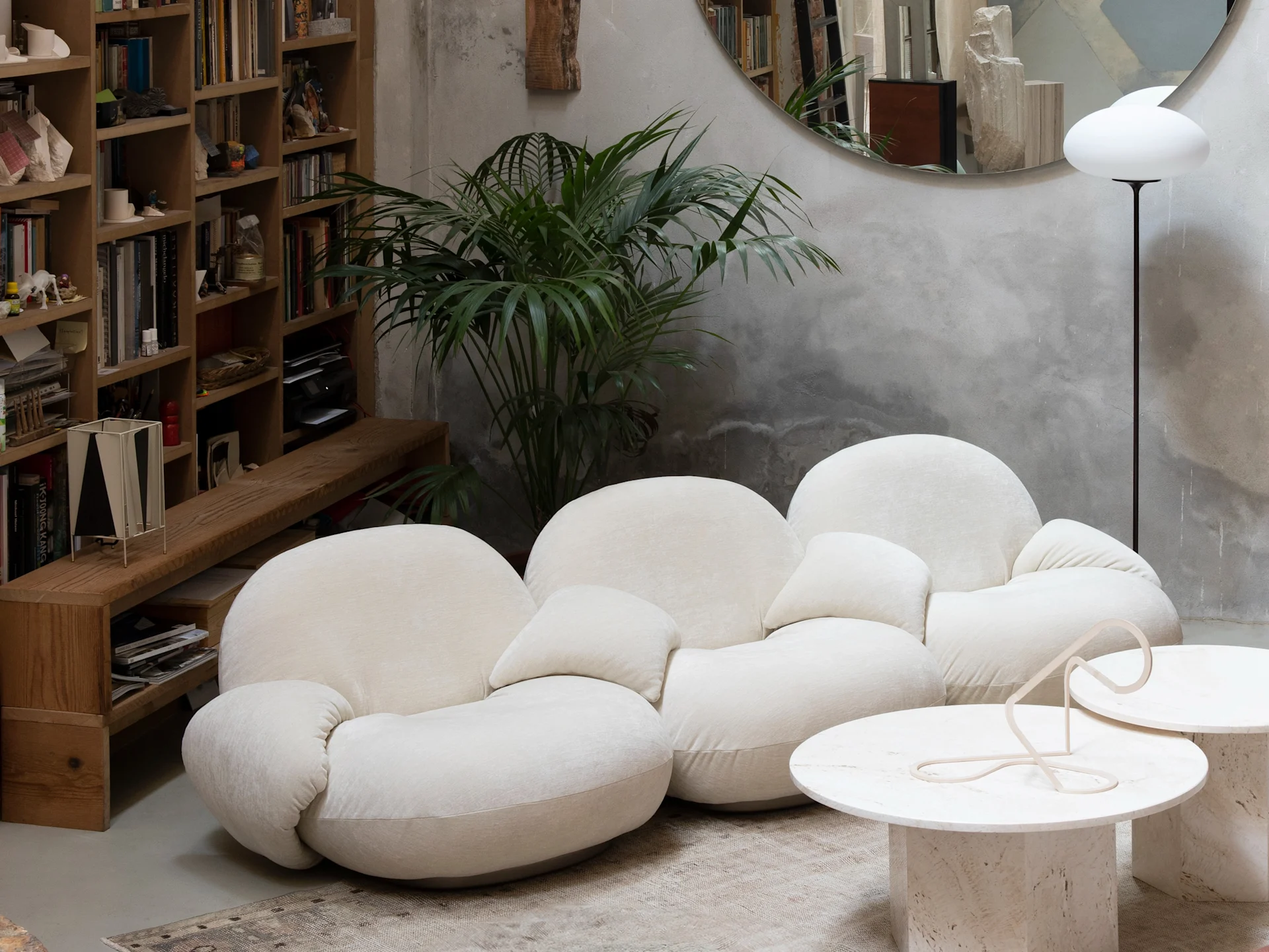 Modern living room with a cream modular sofa, wooden bookshelf, green plant, travertine coffee tables, and a white mushroom-shaped lamp.