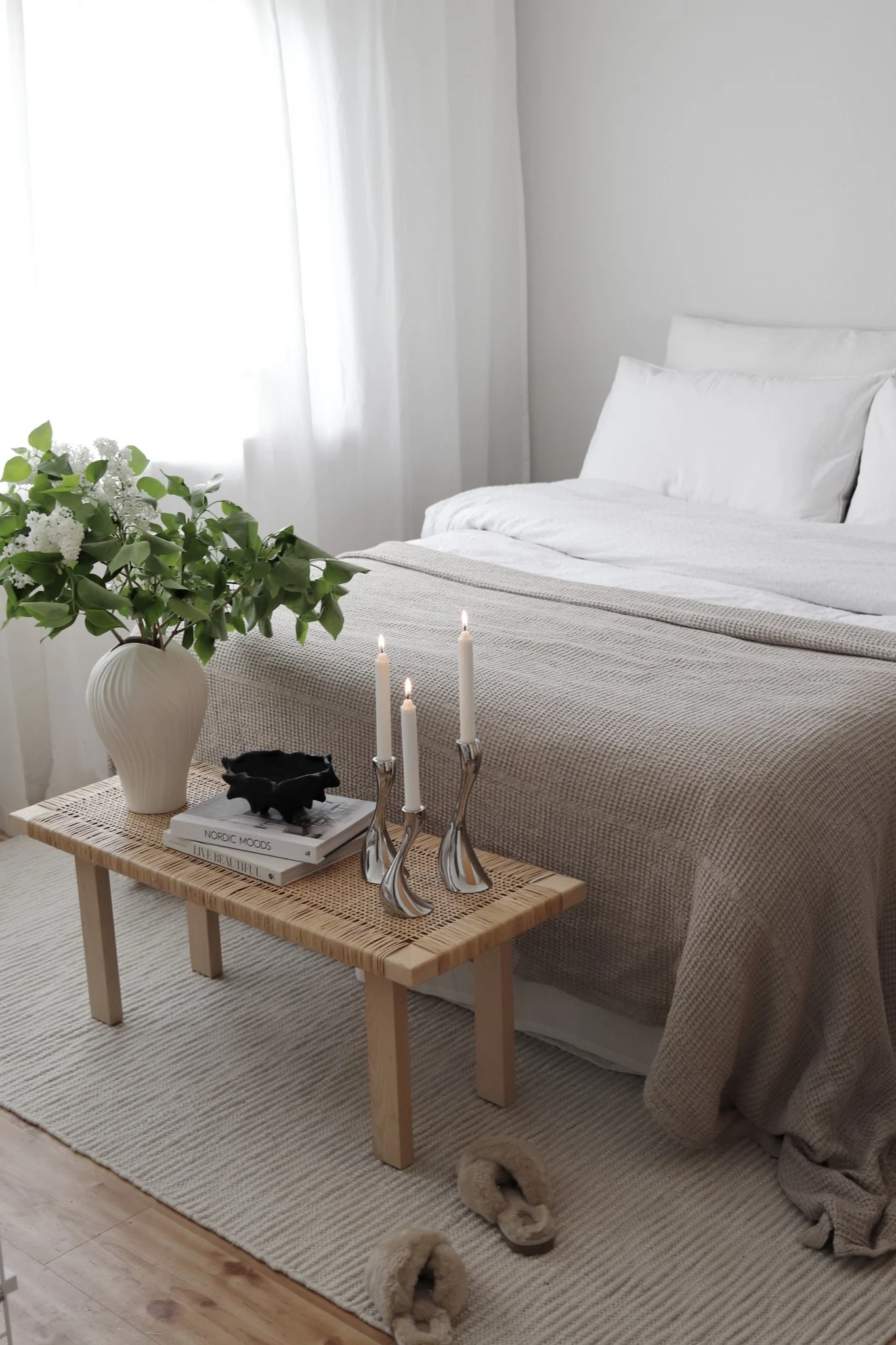 A serene bedroom with a light brown throw on the bed, a wooden bench with white flowers, candles, books, and slippers.