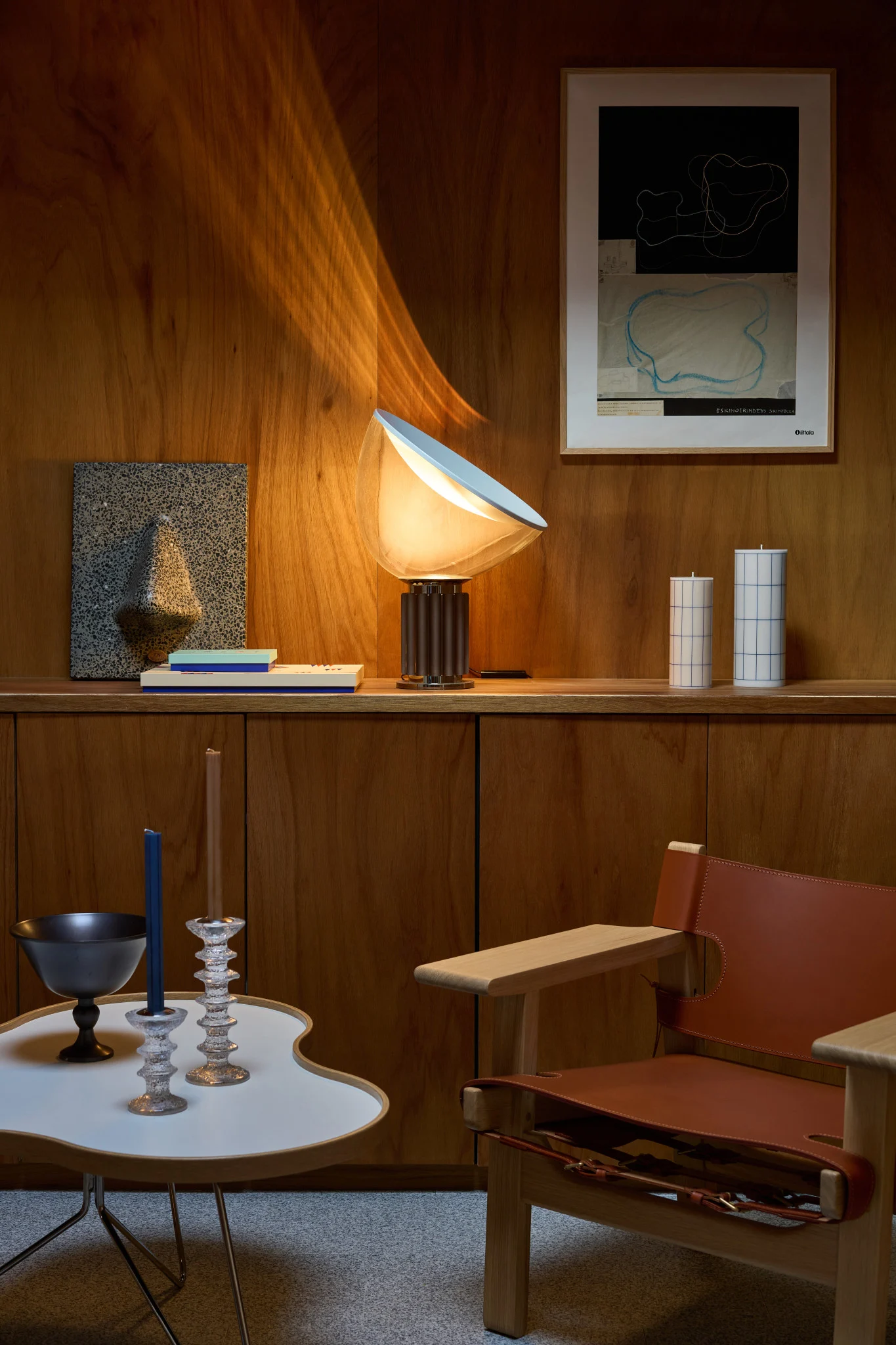 A lamp illuminates a wood-paneled room with shelves, abstract art, candles, a white table, and a leather armchair.