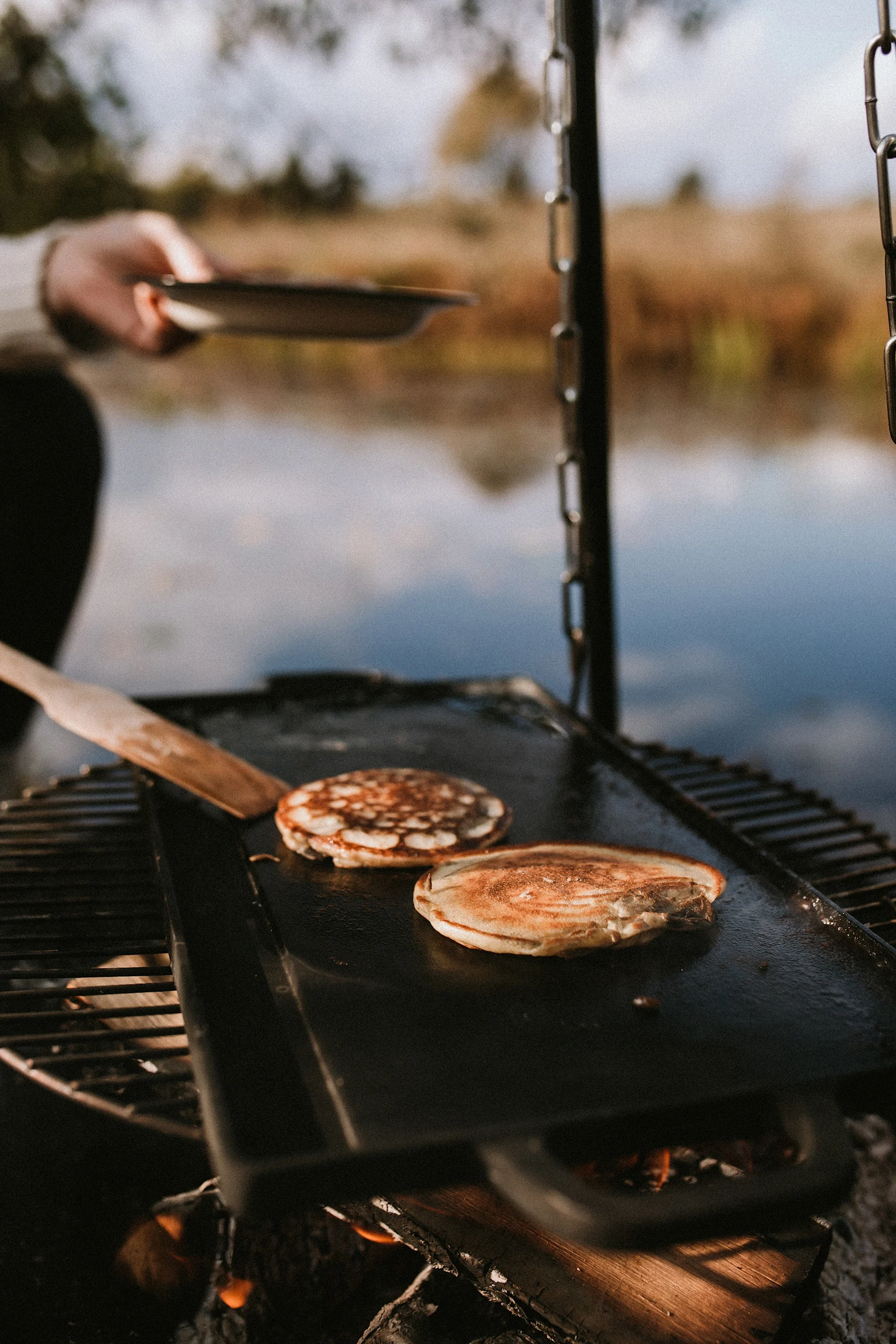 Two pancakes cook on a griddle over a campfire next to a lake, with a hand holding a plate nearby.