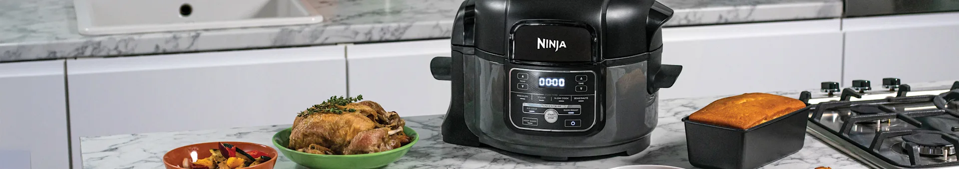 Black multi-cooker on a marble counter, surrounded by roasted chicken, vegetables, and a golden bread loaf.