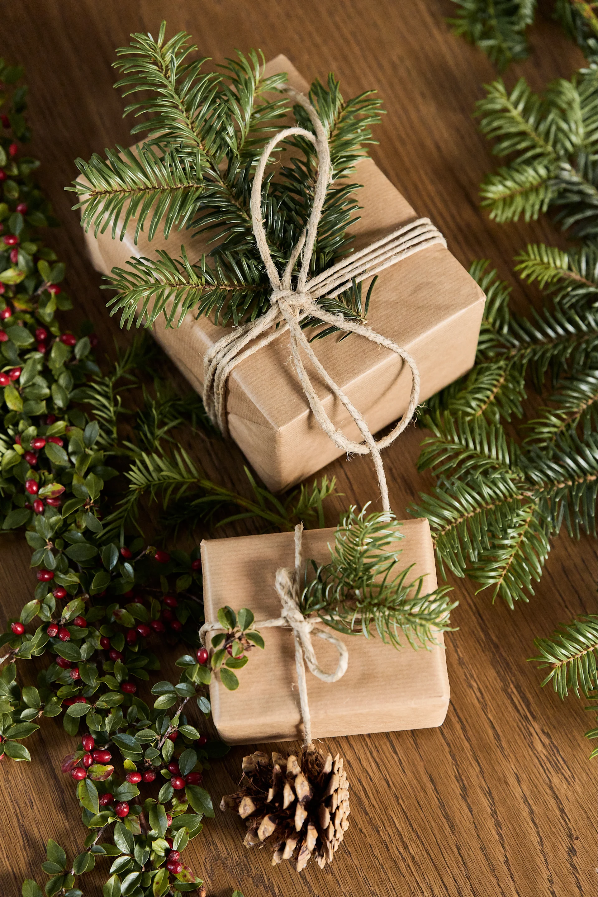 Two brown paper gifts with pine sprigs and twine, red berries, and a pinecone on wood.