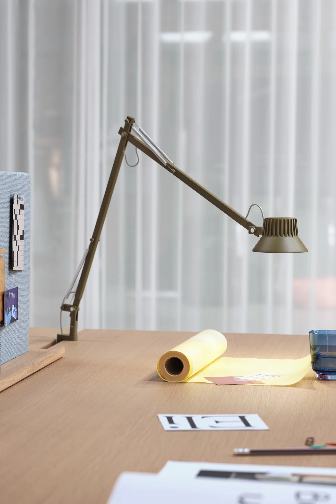 An olive green articulated desk lamp illuminates a light wooden desk with yellow paper, a blue glass, and documents.