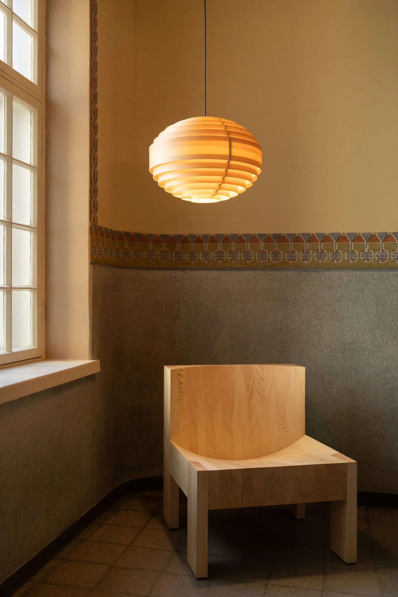 Wooden slatted pendant light above a light wood chair in a room with patterned walls and a window.