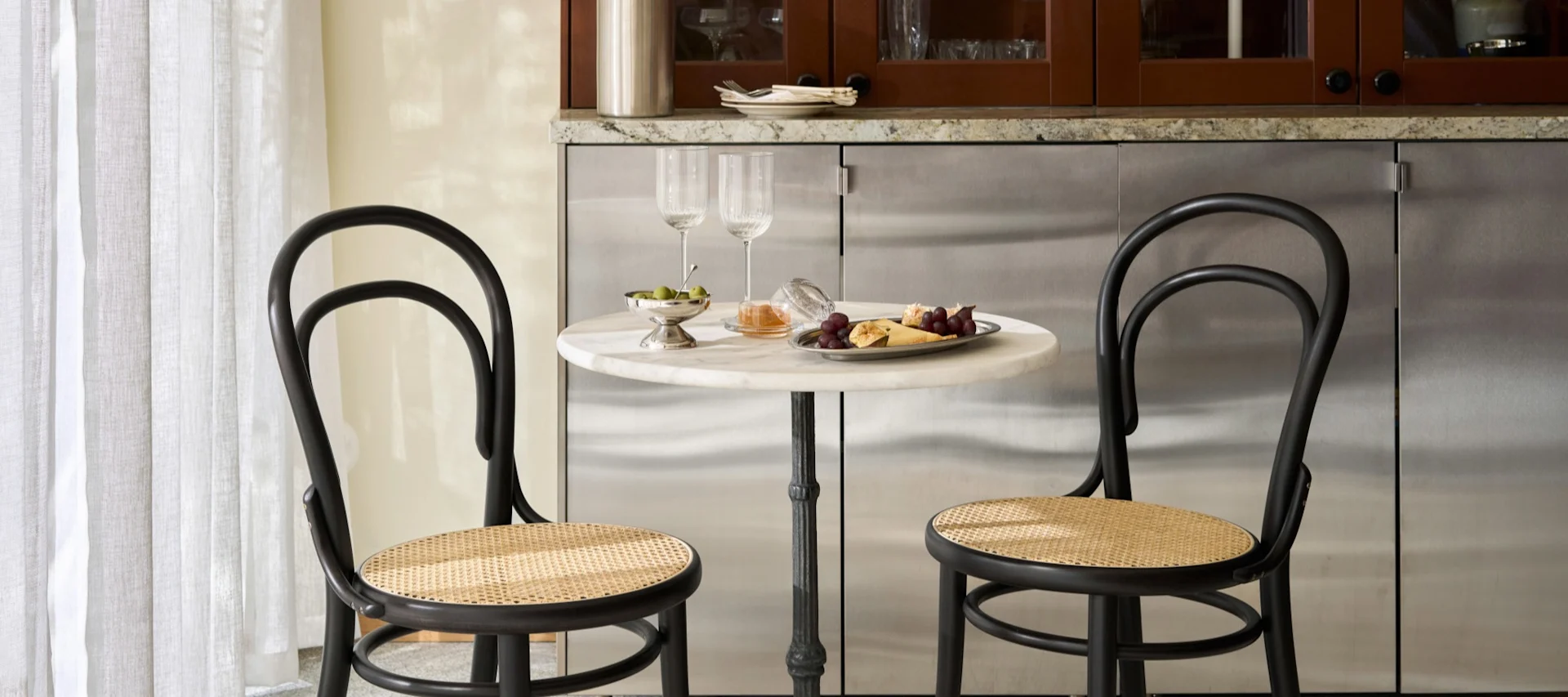 Two black bistro chairs with cane seats at a round marble table set with wine glasses, olives, grapes, and cheese.