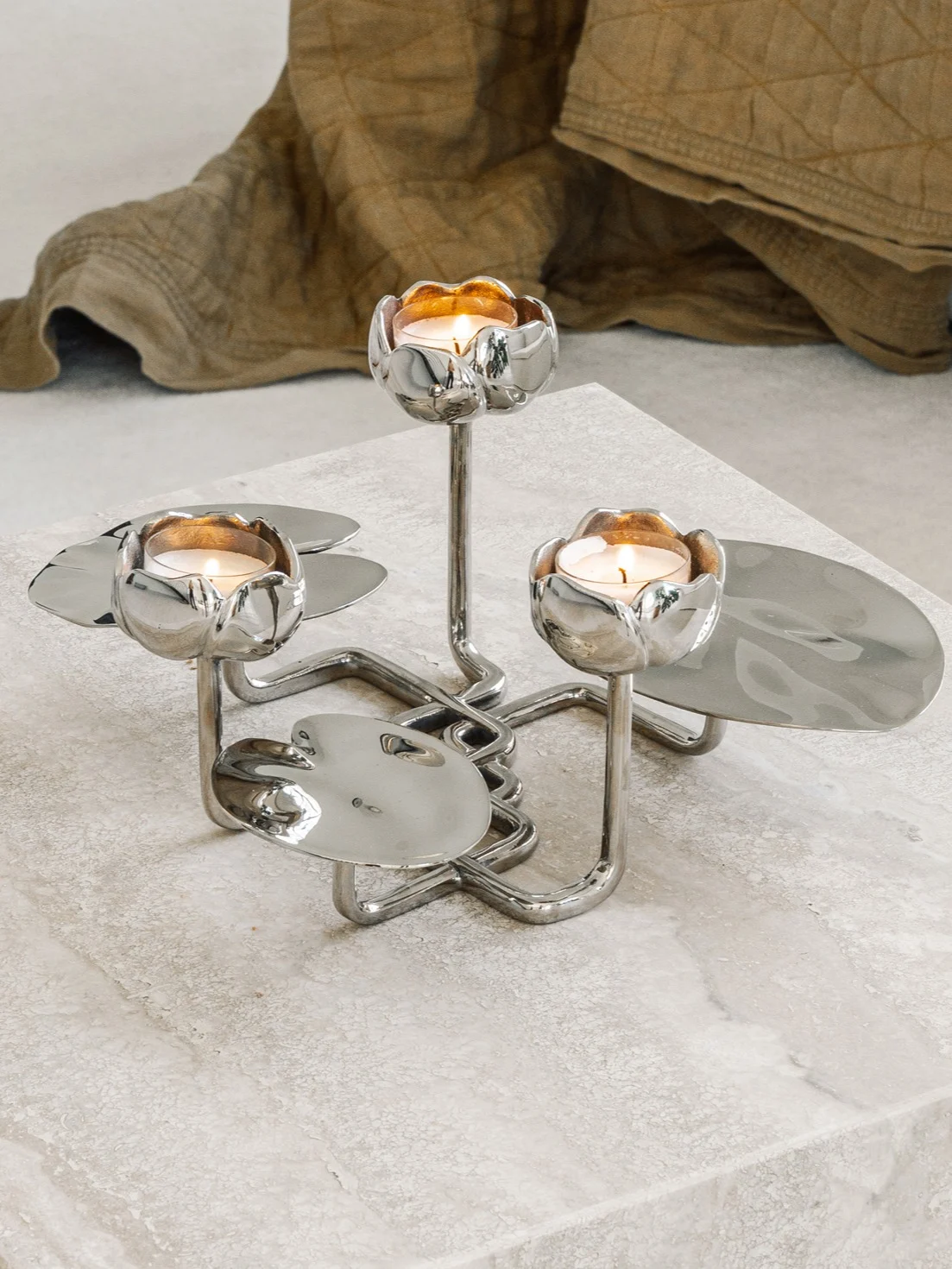 Silver candle holder with three lit candles, designed like water lilies, on a textured surface.