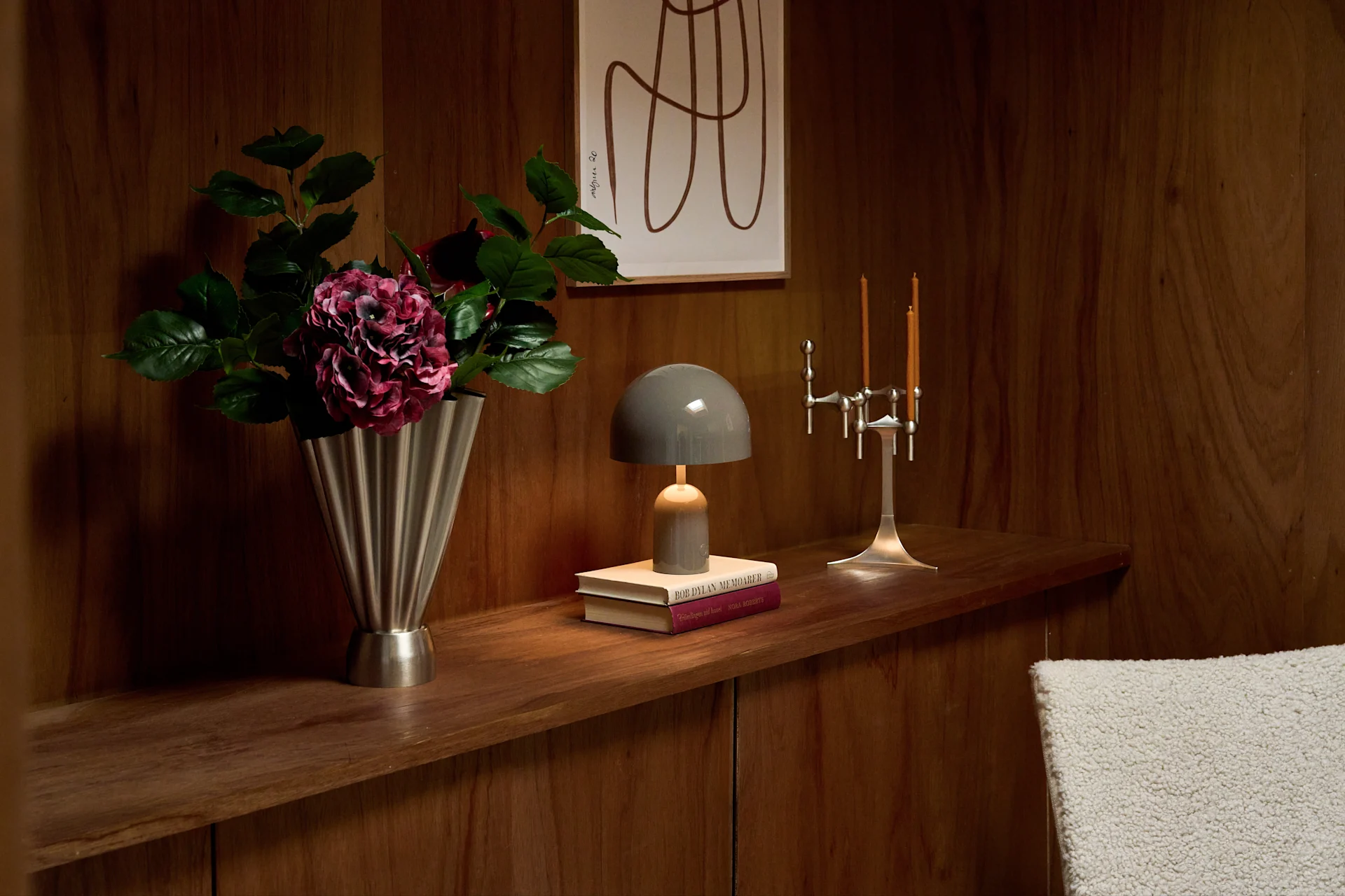 A wooden shelf displays a silver fluted vase with purple flowers, a grey mushroom lamp on books, and a silver candelabra with two lit candles.