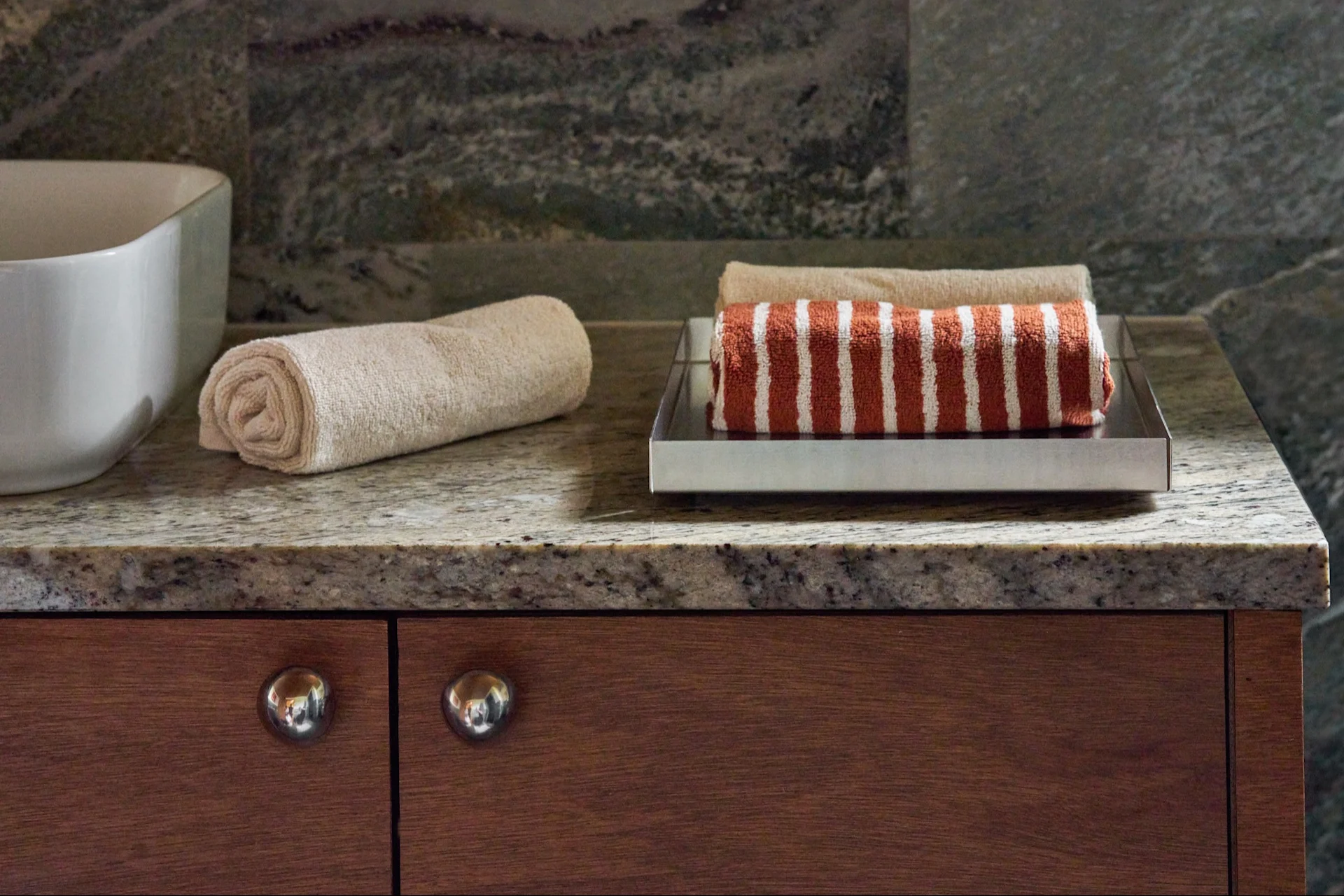 Rolled beige and striped rust-colored towels on a silver tray on a granite countertop, next to a white sink basin and wooden cabinets.
