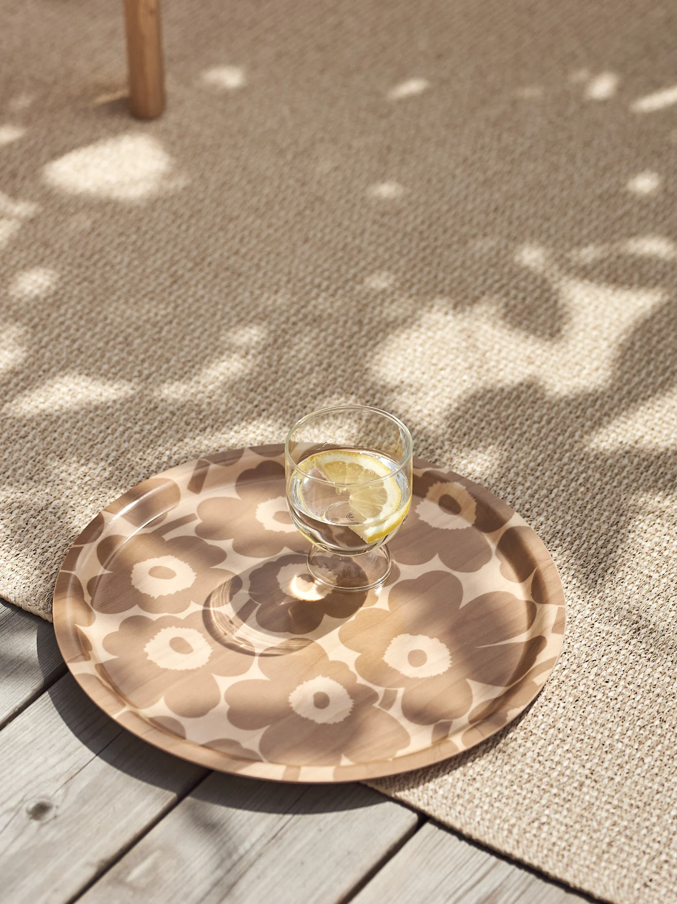 Glass of lemon water on a beige floral patterned tray on a textured rug with dappled sunlight.