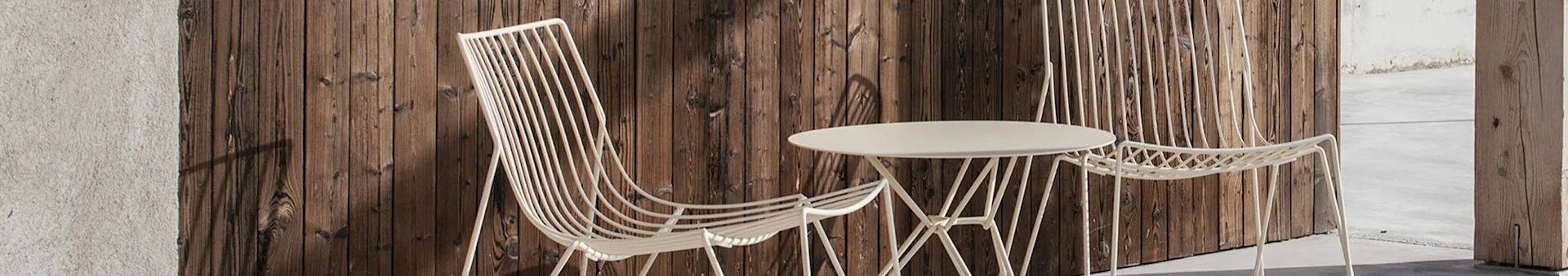 Two light-colored, slatted metal outdoor chairs and a matching round table with a geometric base, against a dark wooden background.