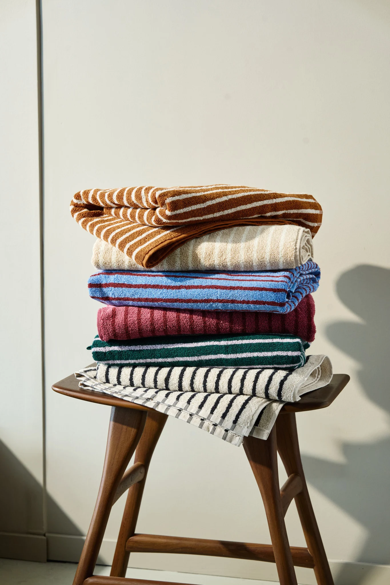 A stack of colorful striped and solid towels in brown, beige, blue, maroon, green, and black on a wooden stool.
