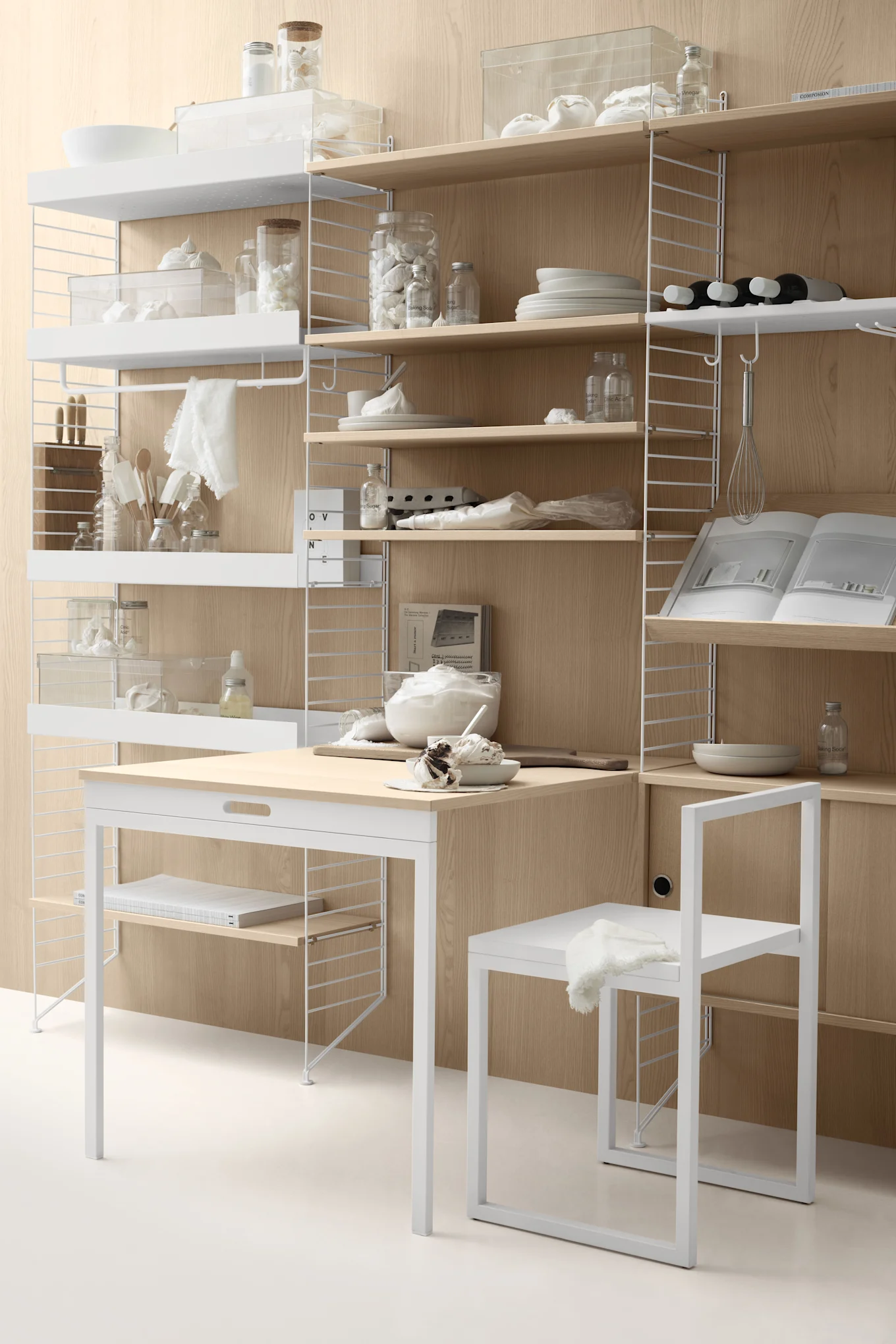 A kitchen setup with white modular shelving on a wood-paneled wall, featuring baking ingredients, utensils, a light wood desk, and a white chair.