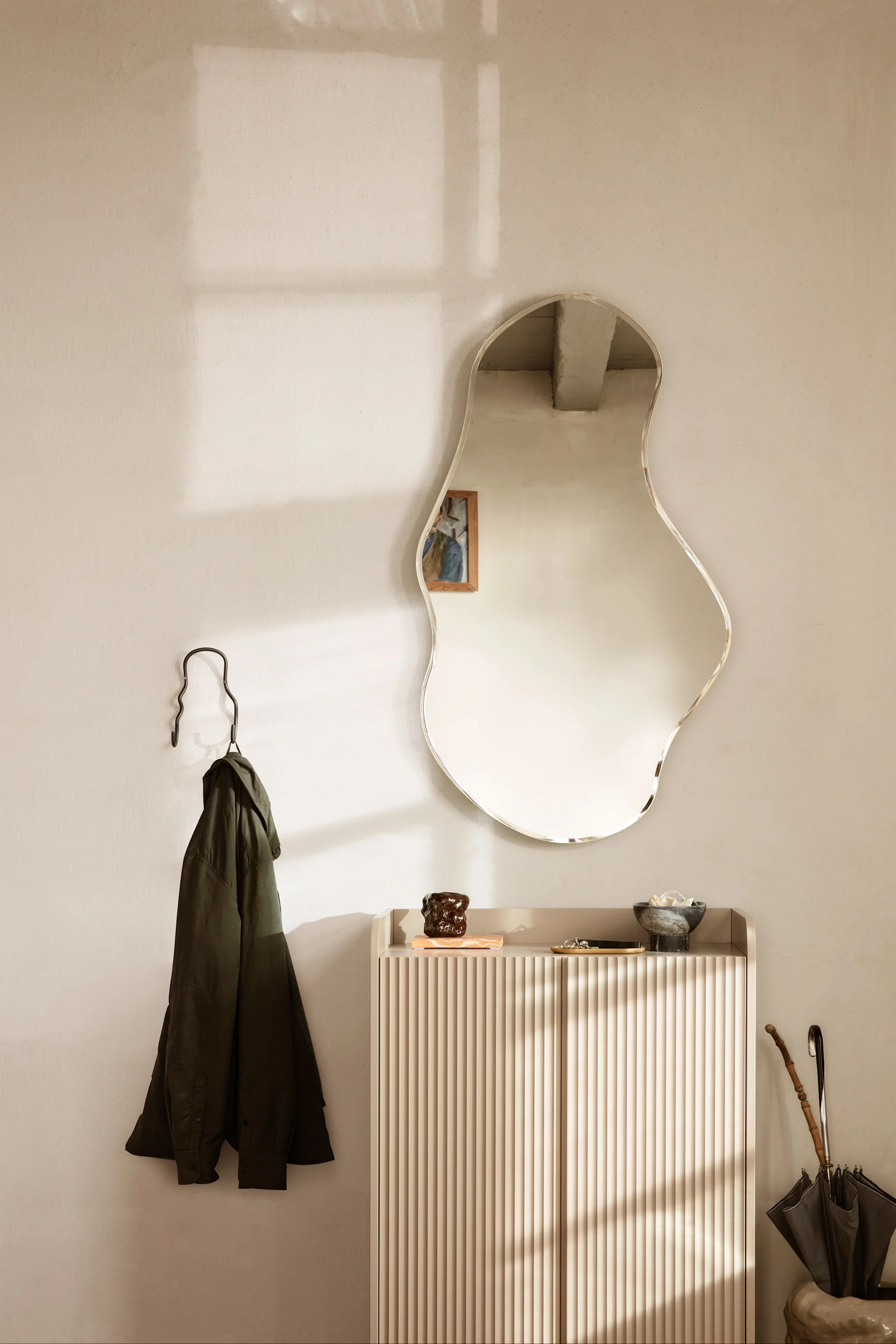 An entryway features an organic-shaped mirror above a fluted console, a green jacket on a hook, and an umbrella, with sunlight.
