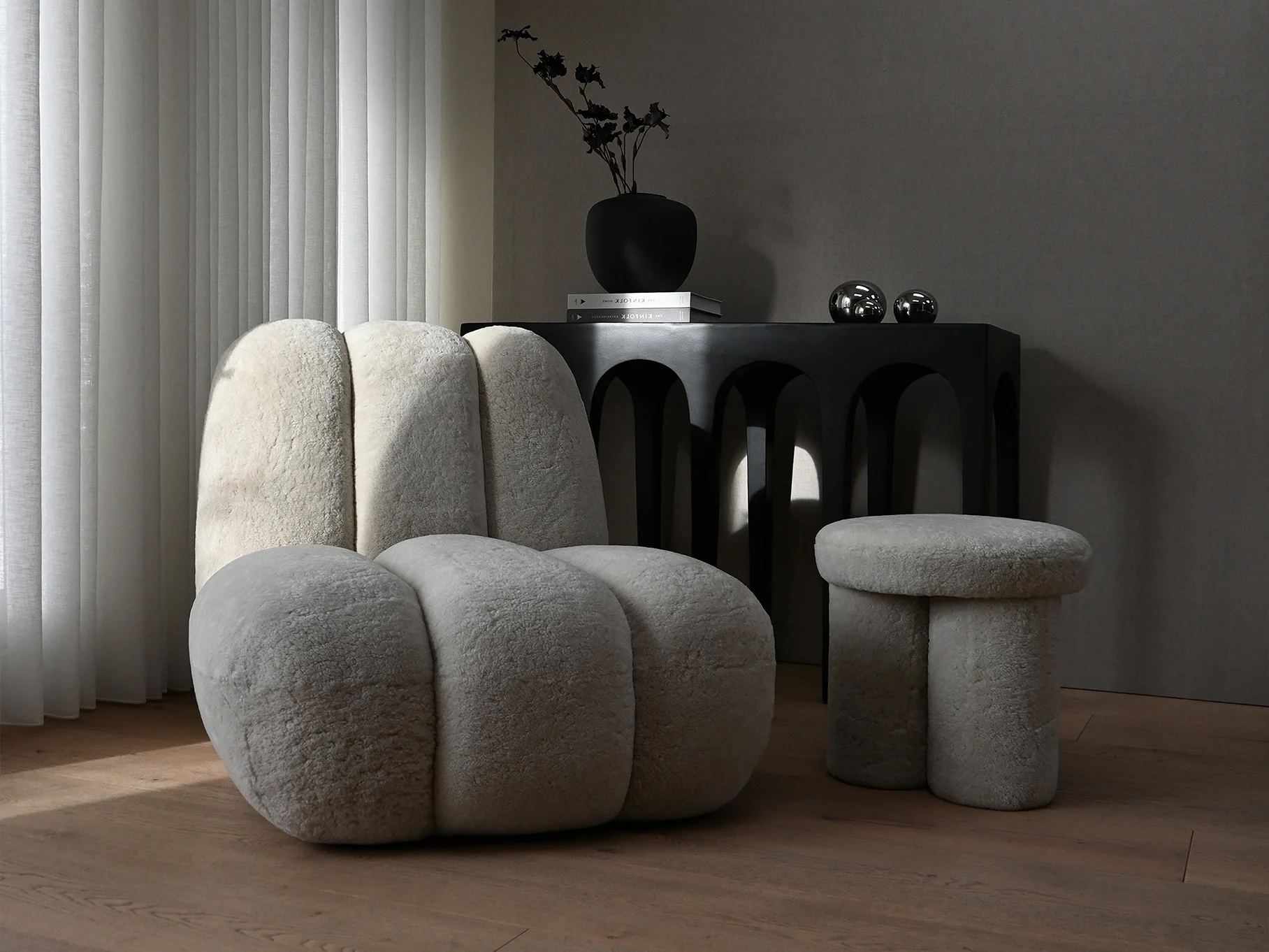 A light-colored, fluffy armchair and a matching three-legged stool in a modern room with a dark console table and white blinds.