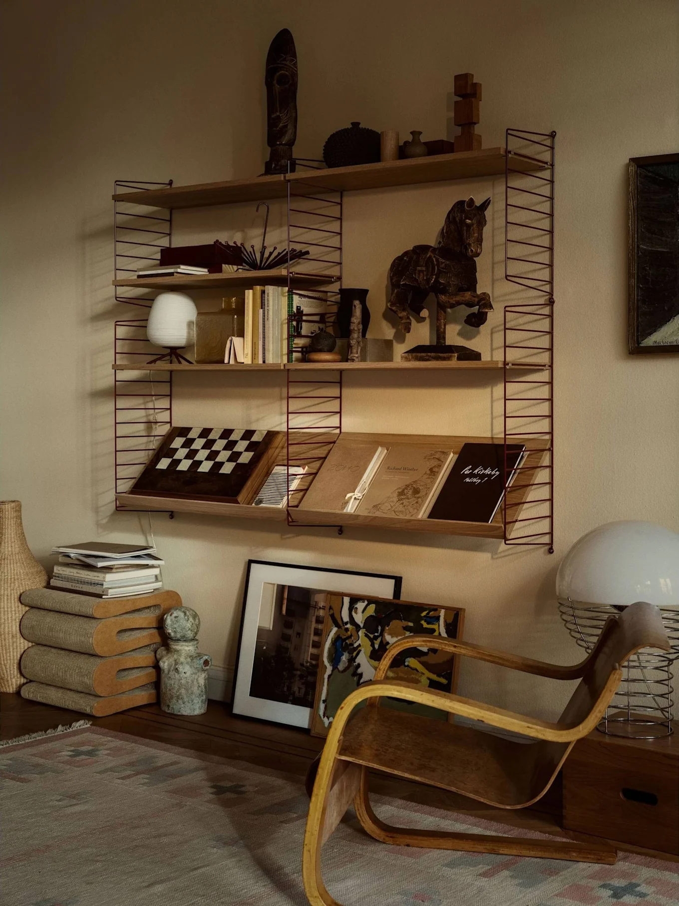 A cozy room featuring a shelving unit filled with books, art, and decor, beside a wooden bentwood armchair and a large white lamp.