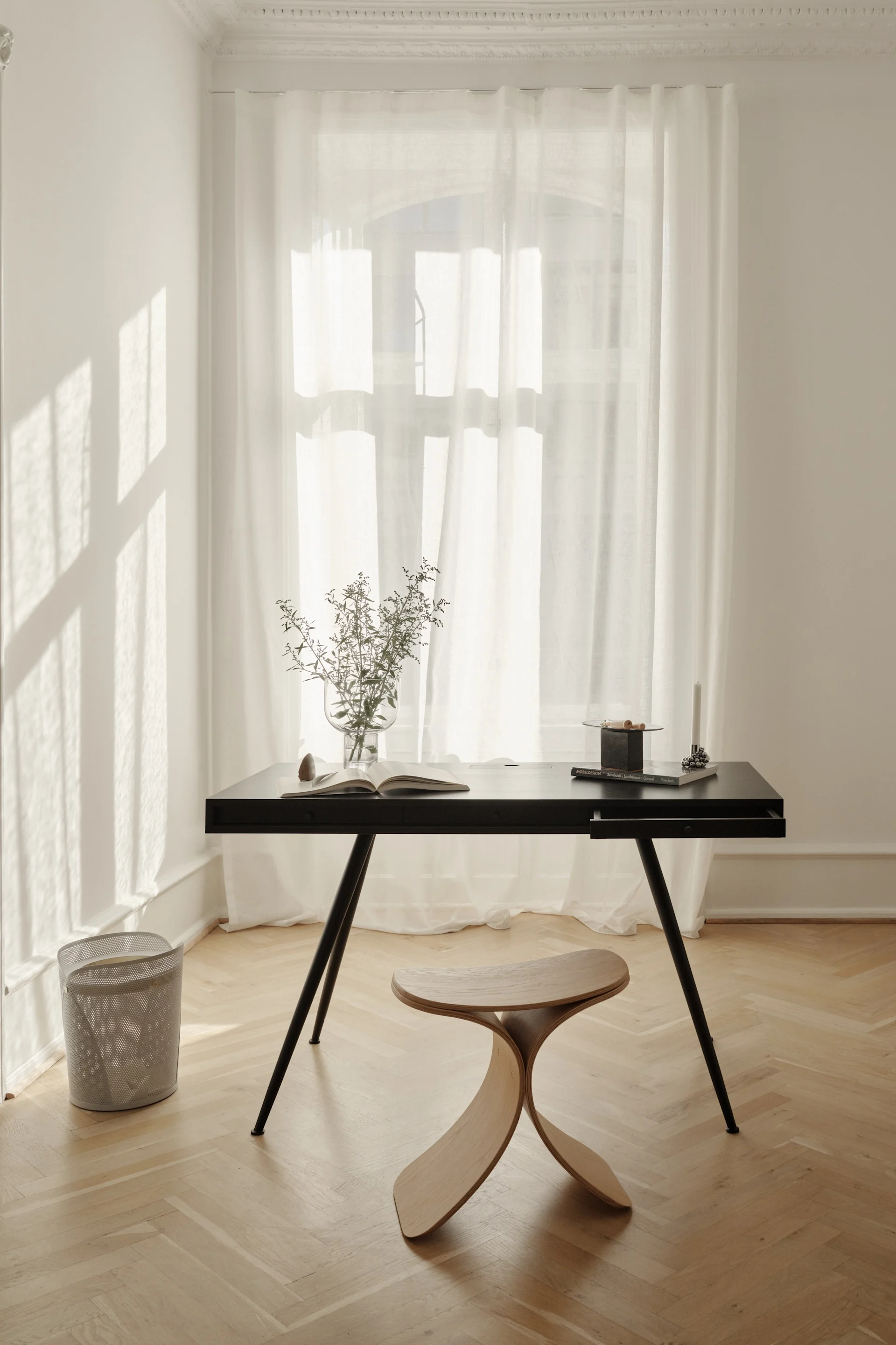 A minimalist home office with a black desk, a curved wooden stool, a vase of foliage, and an open book by a window with sheer curtains.