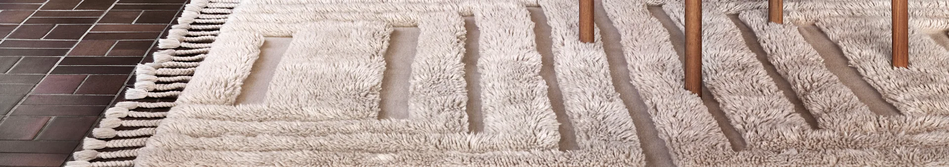 Close-up of a light beige shaggy rug with a geometric pattern and tassels on dark brown tiled floor. Wooden furniture legs are on the rug.