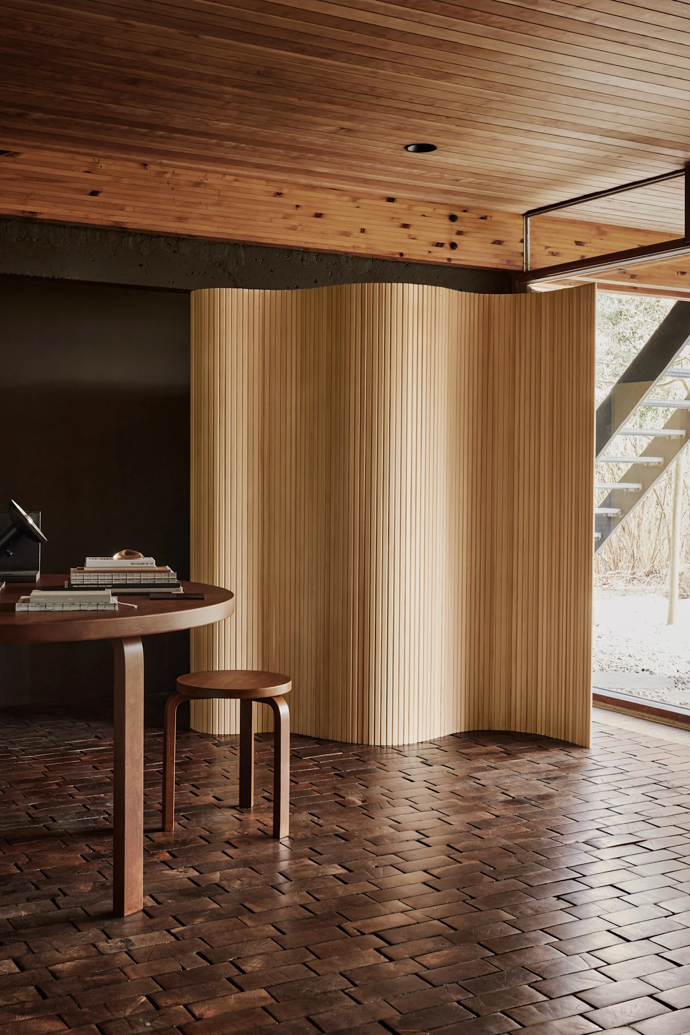Modern room with a round wooden table, a small stool, and a wavy wooden screen. Dark wood block floor, light wood slat ceiling, and a window to an outdoor staircase.