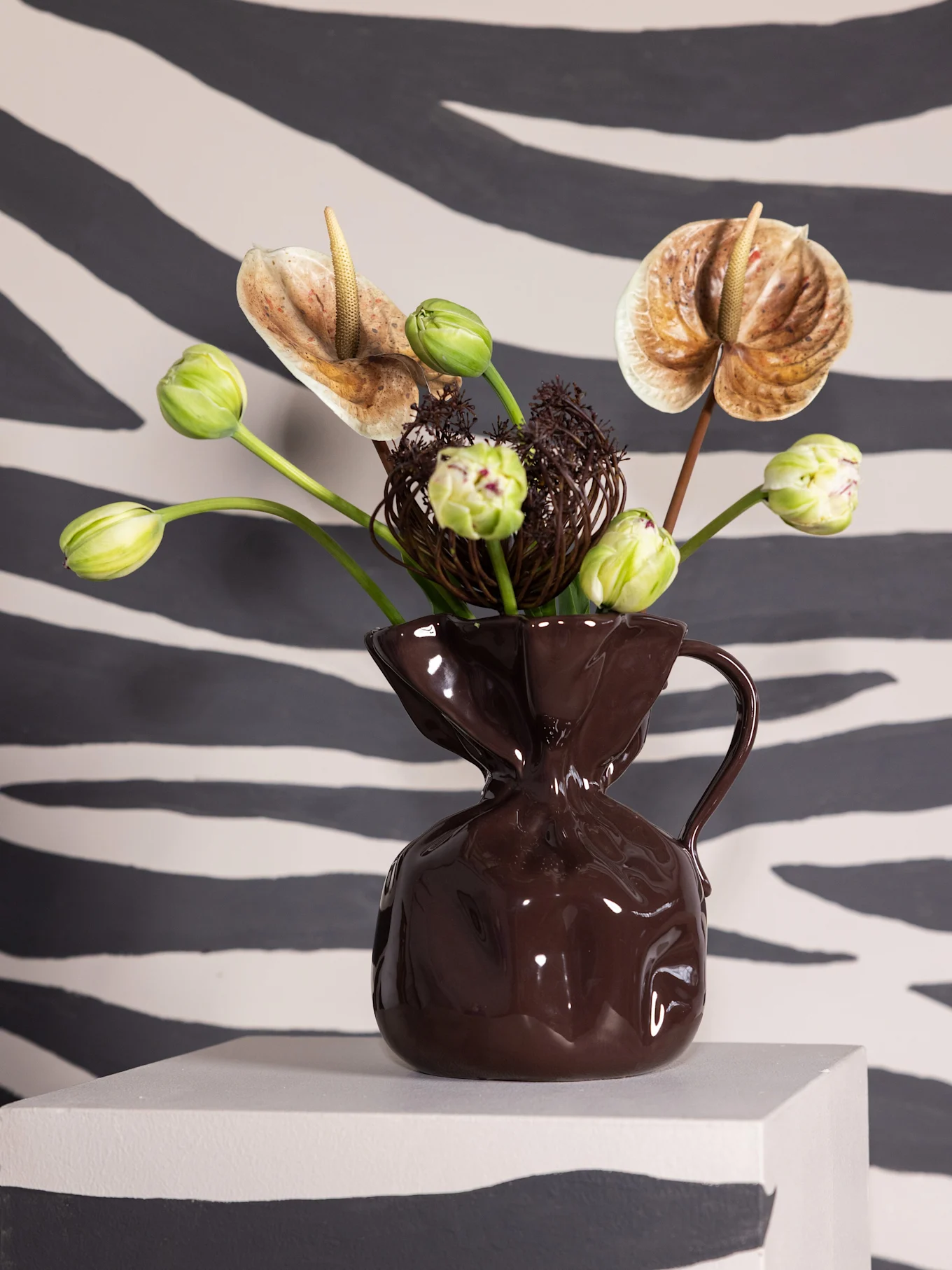 Floral arrangement in a dark brown vase with anthuriums and green tulips on a grey pedestal against a zebra-striped background.