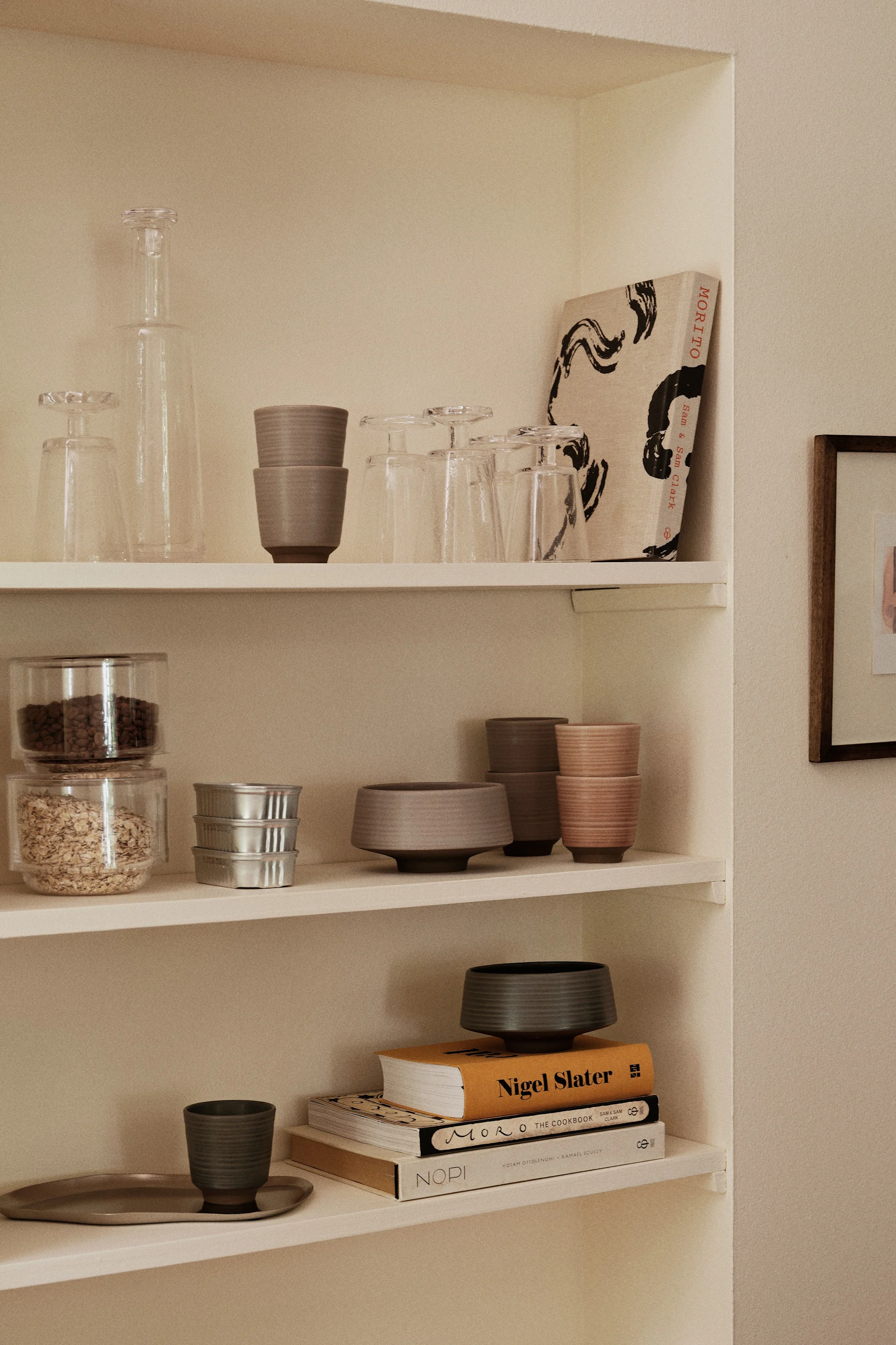 Built-in shelves display clear glass items, stacked ceramic cups and bowls in neutral tones, food containers, silver tins, and cookbooks.