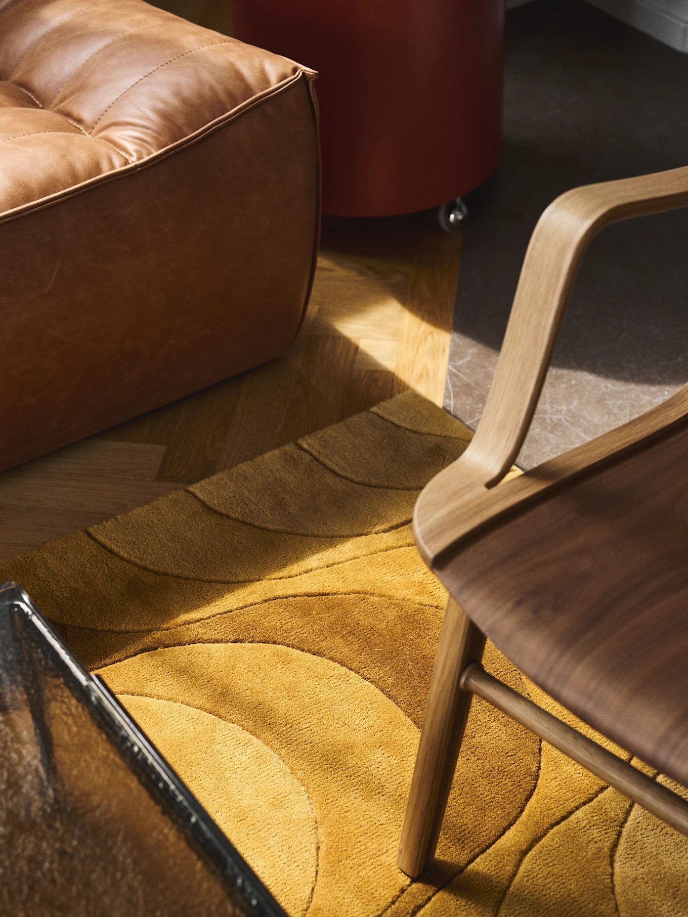 A brown tufted leather ottoman rests beside a golden rug with wavy patterns, a curved wooden chair, and a glass table in a sunlit room.
