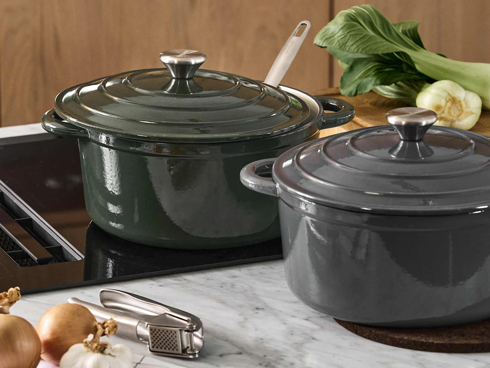 Two lidded cast iron pots, one dark green and one dark grey, on a kitchen countertop with bok choy, onions, and a garlic press.