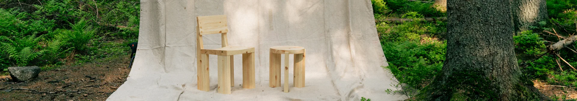 A light wood chair and a matching round stool on a canvas backdrop in a forest setting.