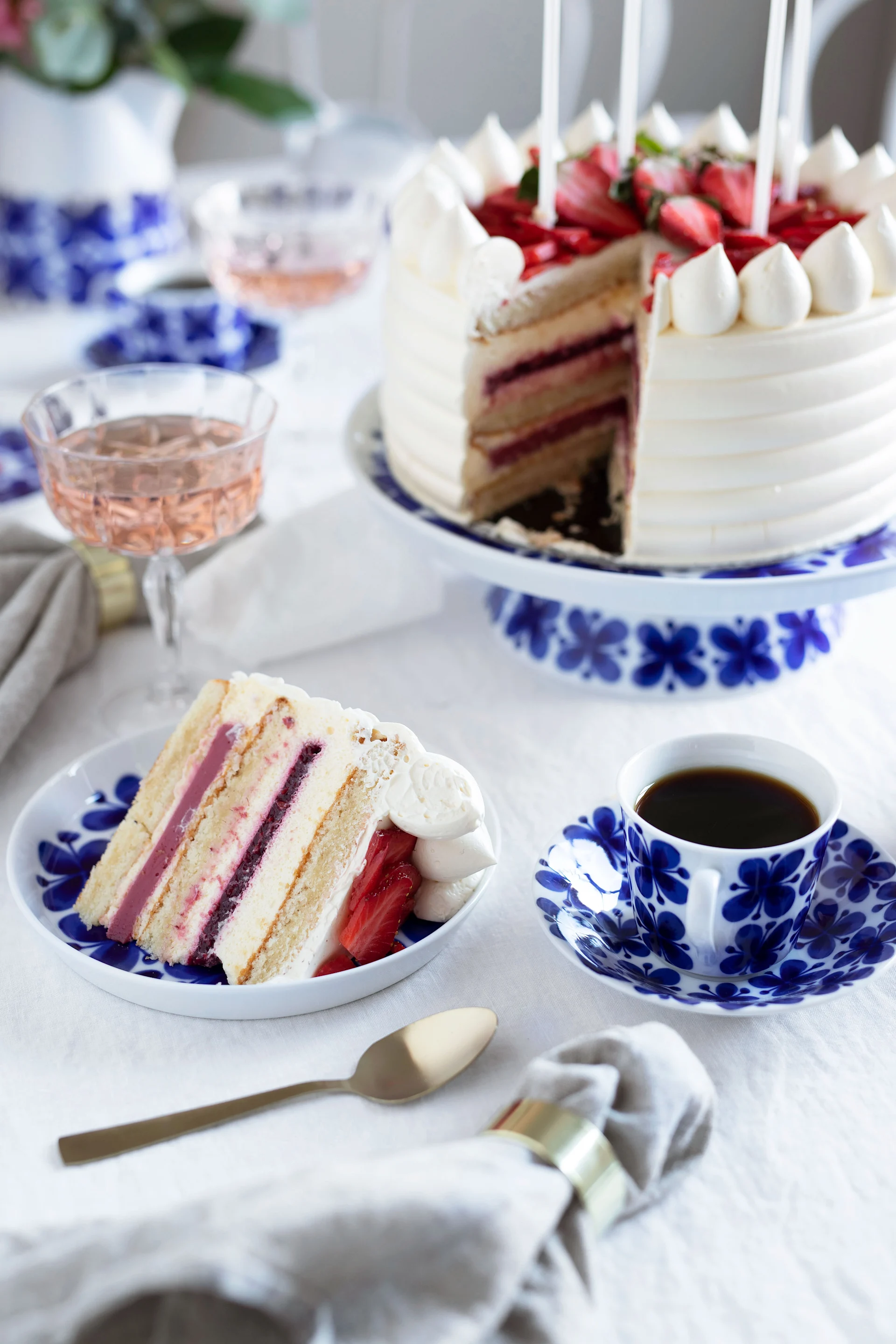 Strawberry cake with a slice cut out, served on blue and white patterned dinnerware with coffee and rosé.