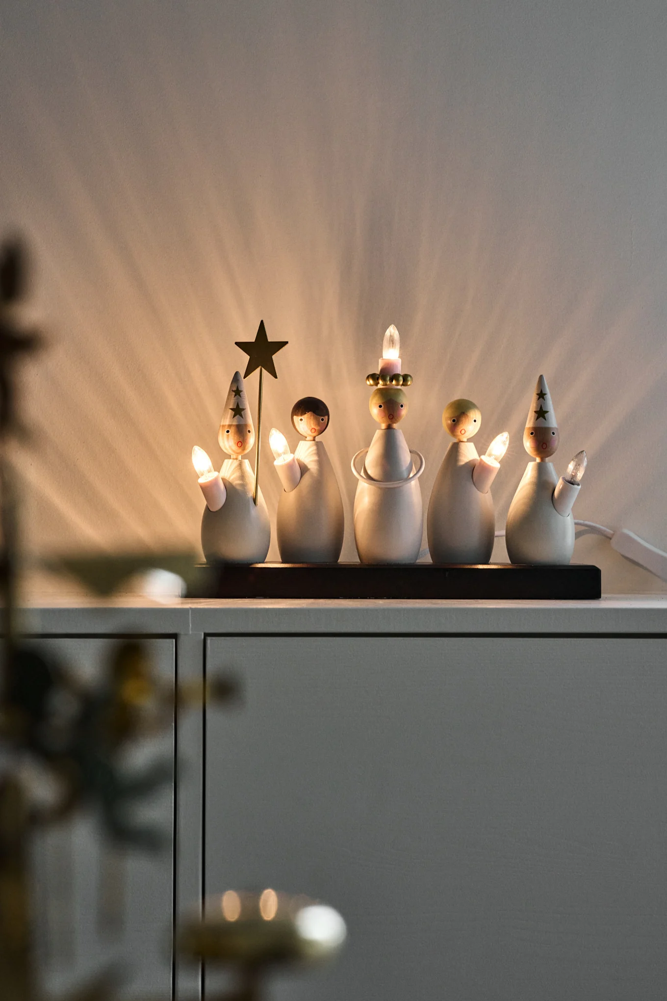 Five minimalist wooden Christmas figures, some with star hats, holding candle lights on a cabinet, casting warm light.