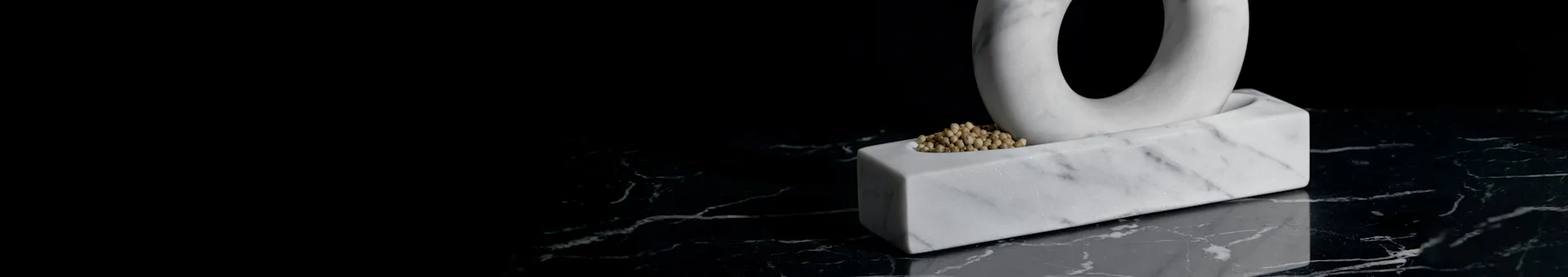 White marble sculpture featuring an abstract ring shape emerging from a rectangular base filled with small beads, on a dark marble surface.