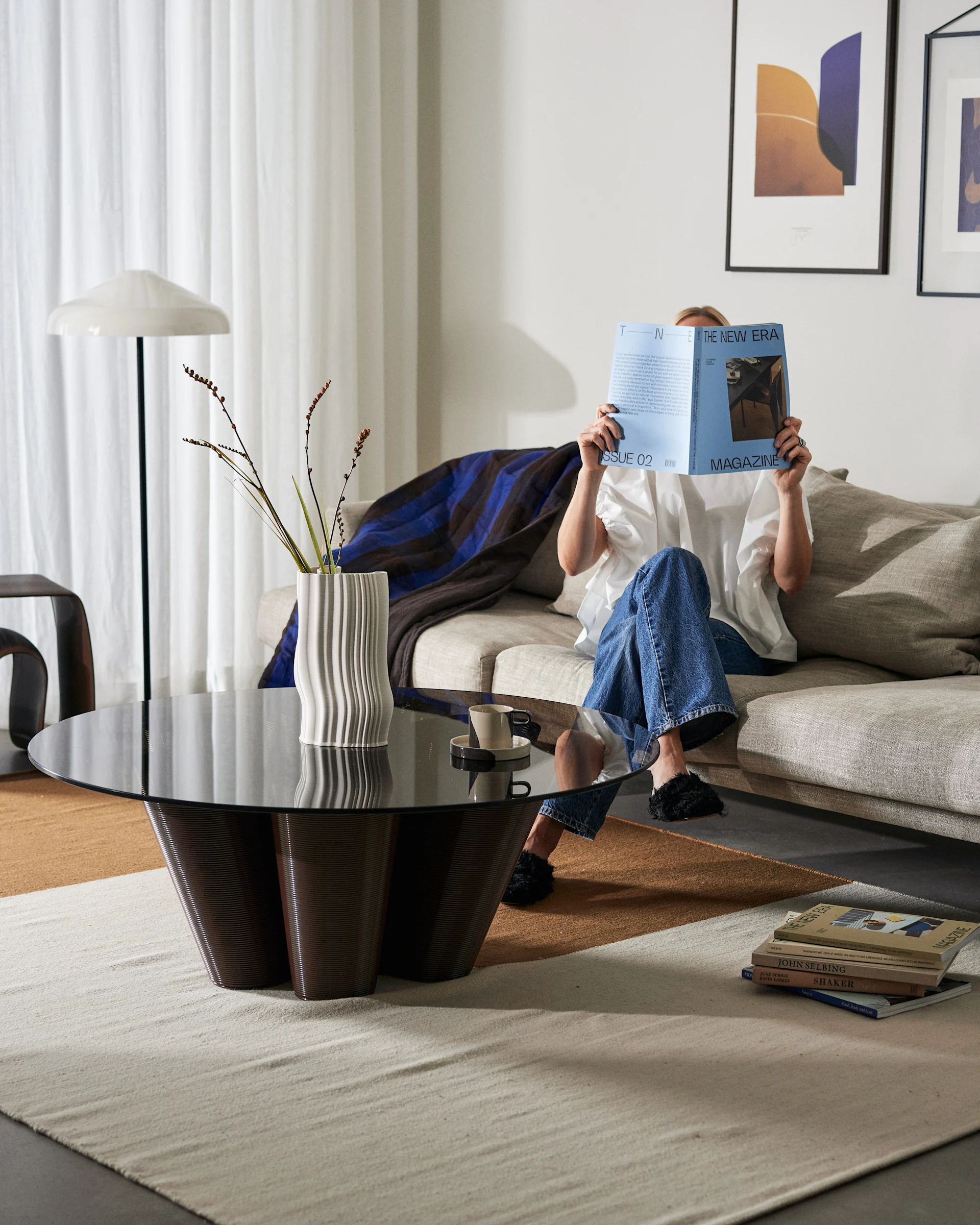 A person in blue jeans reads a blue magazine on a light sofa in a minimalist living room with a dark coffee table.