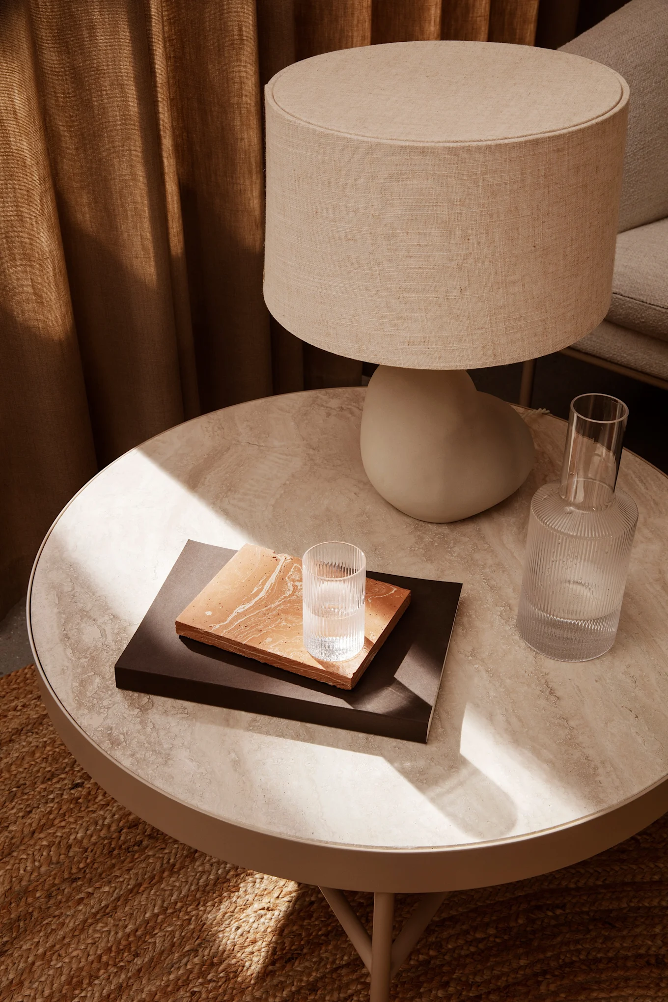 A round marble-patterned side table with a fabric lamp, two nested rectangular trays, a glass of water, and a ribbed glass carafe, all on a woven rug.
