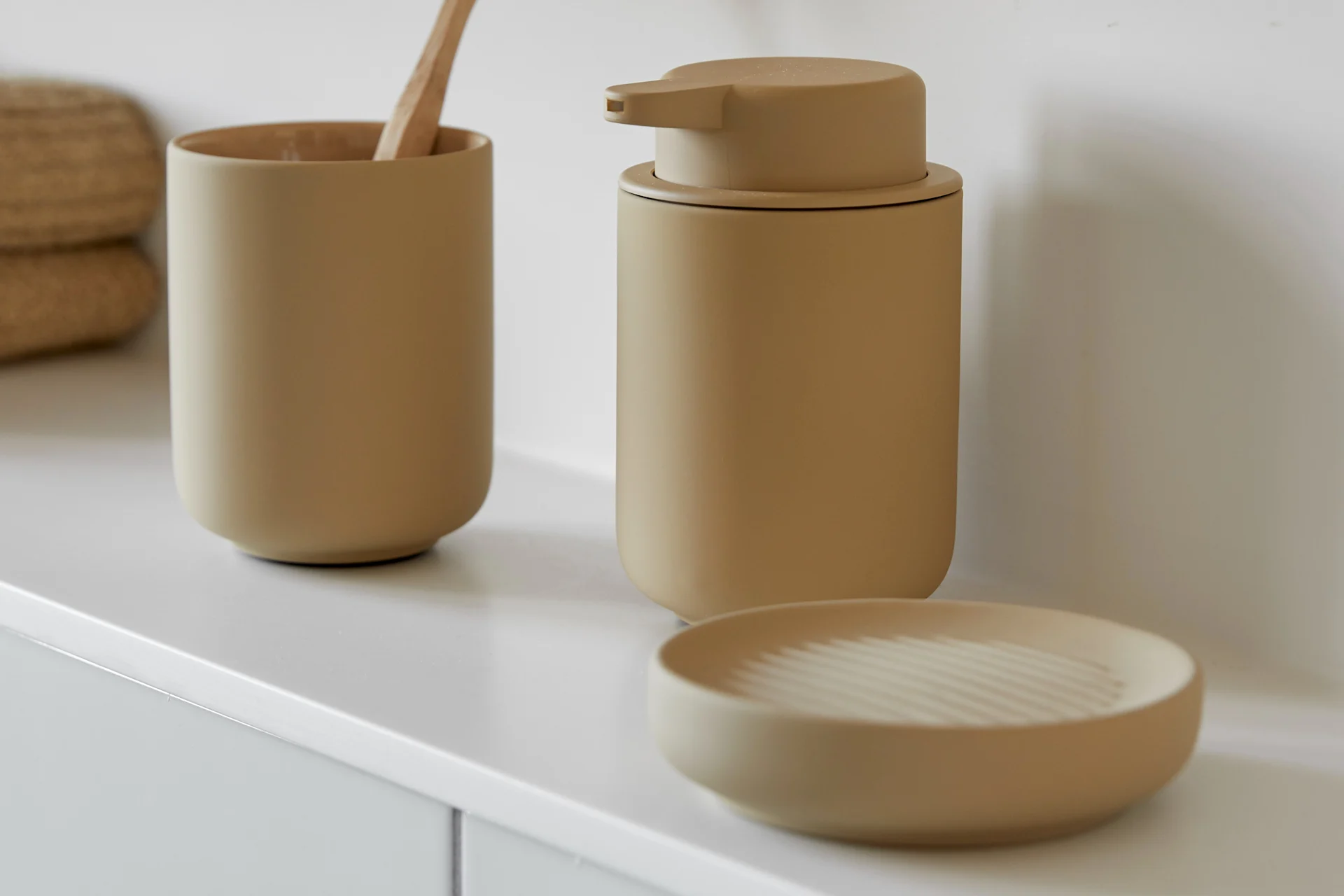 A light brown toothbrush holder with a wooden toothbrush, a matching soap dispenser, and a soap dish on a white counter.