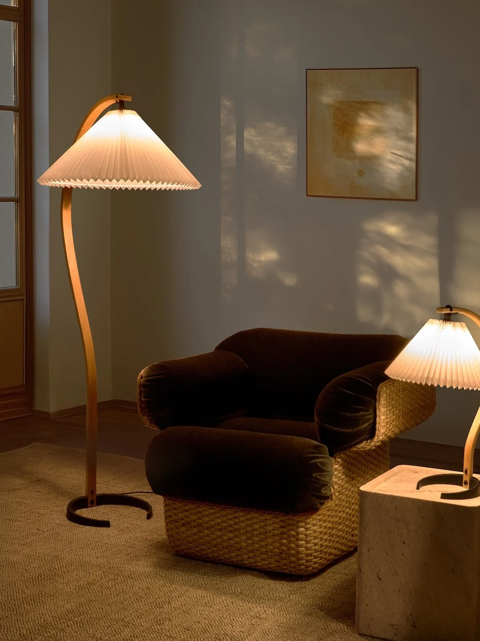 Cozy room with a curved floor lamp, dark armchair with woven base, and a smaller table lamp. Warm light creates shadows.