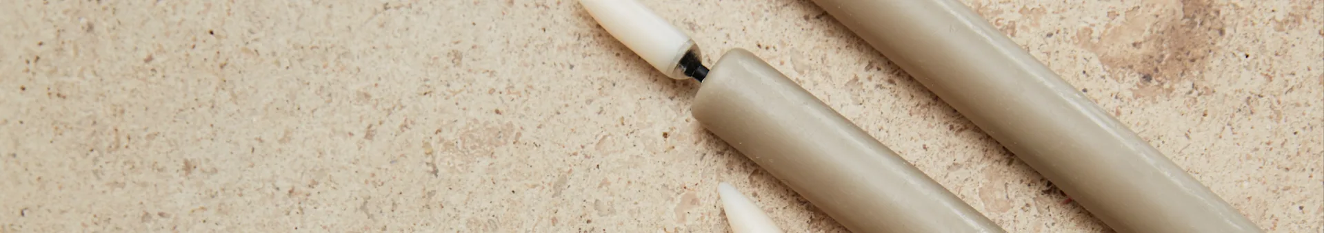 Three unlit beige candles on a speckled light brown surface. One candle is broken, showing its black wick.