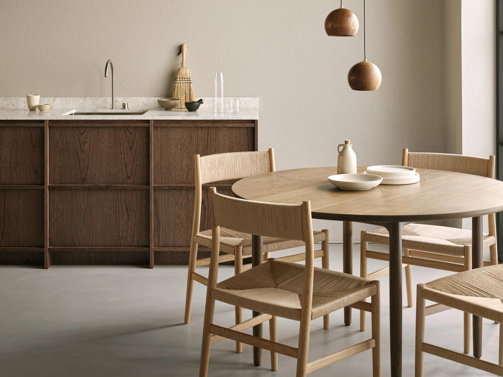 A modern dining area and kitchen. A round wooden table with woven chairs and two spherical pendant lights. Dark wood cabinets line a light stone counter.