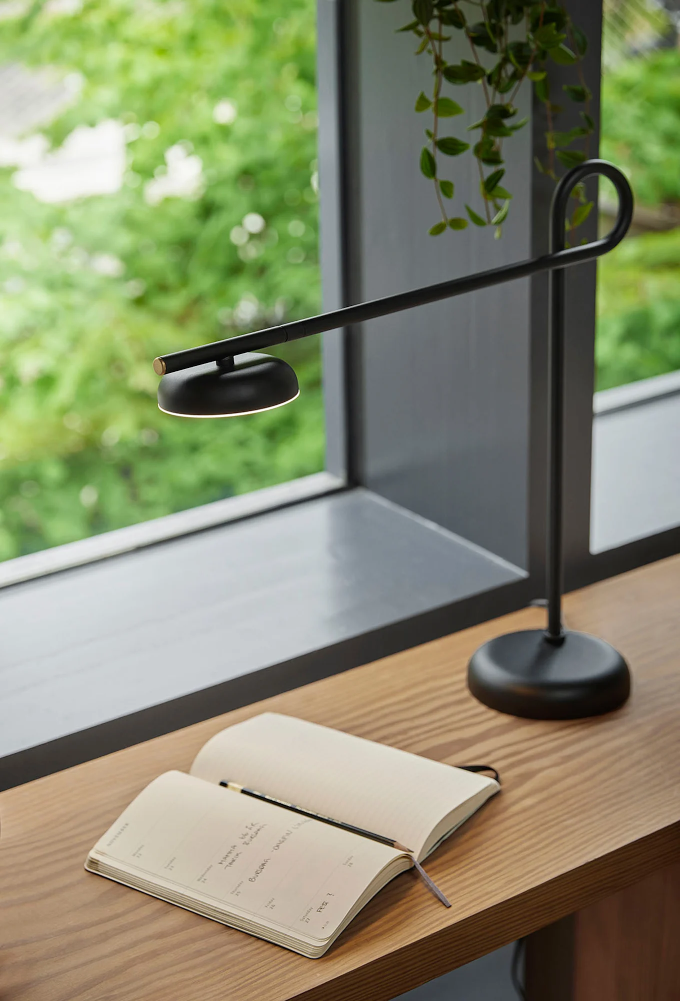 A modern black desk lamp illuminates an open notebook and pencil on a wooden desk next to a window with green foliage.