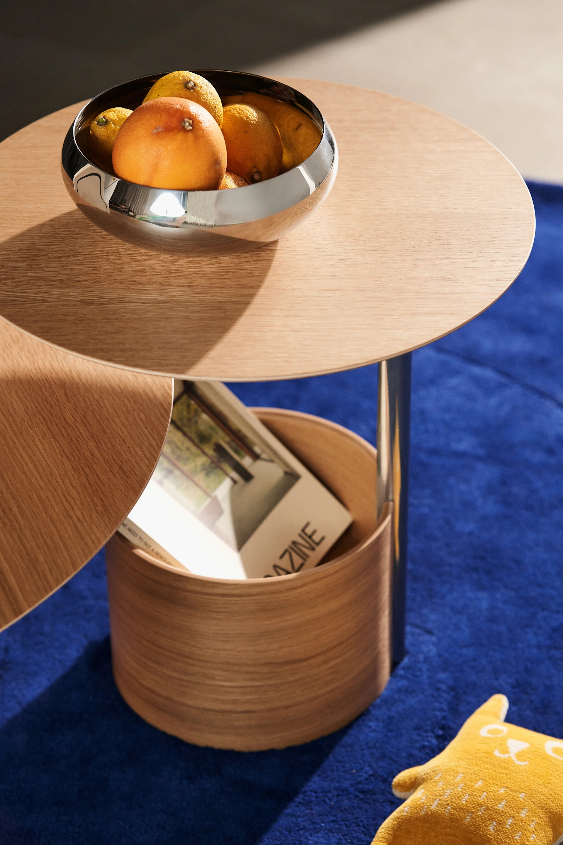 A round wooden coffee table with a chrome bowl of oranges, a lower storage unit with a magazine, and a yellow cat cushion on a blue rug.