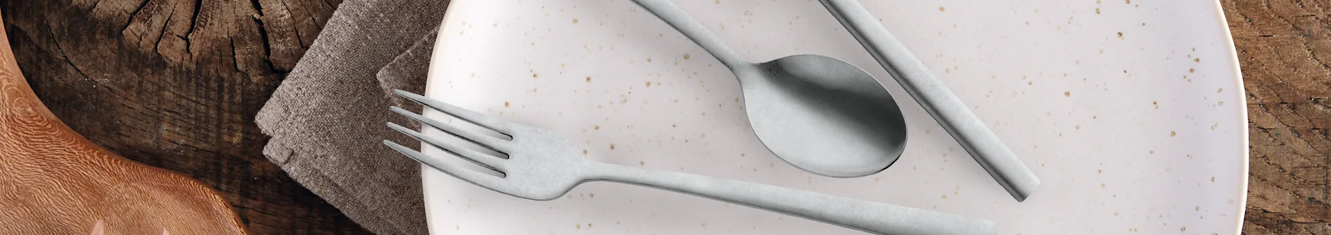 Close-up of a dining table setting with a fork, spoon, and knife on a speckled white plate, next to a textured napkin and a wooden surface.