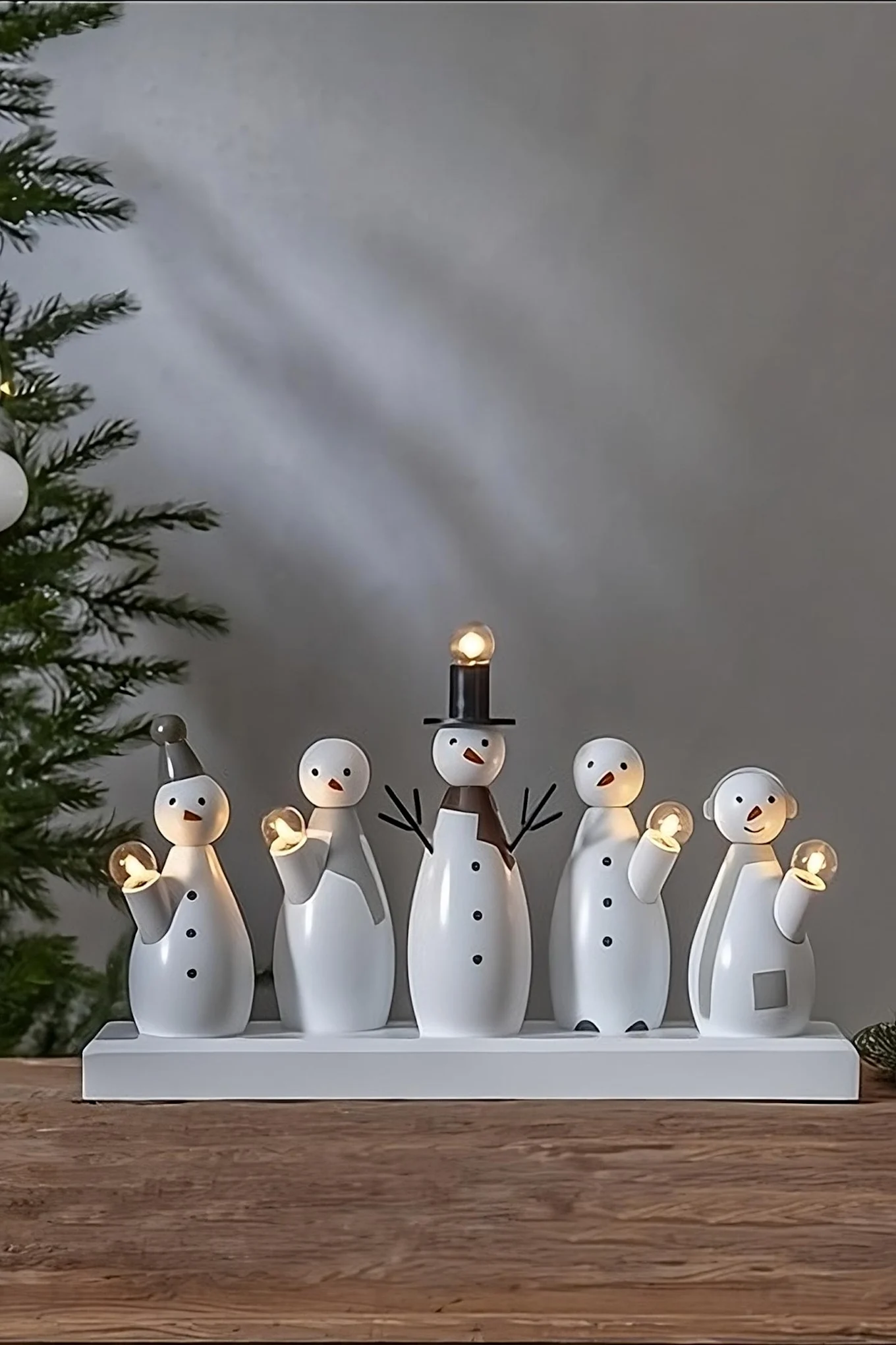 Five white wooden snowman figures with glowing lights on a white base, next to a Christmas tree.