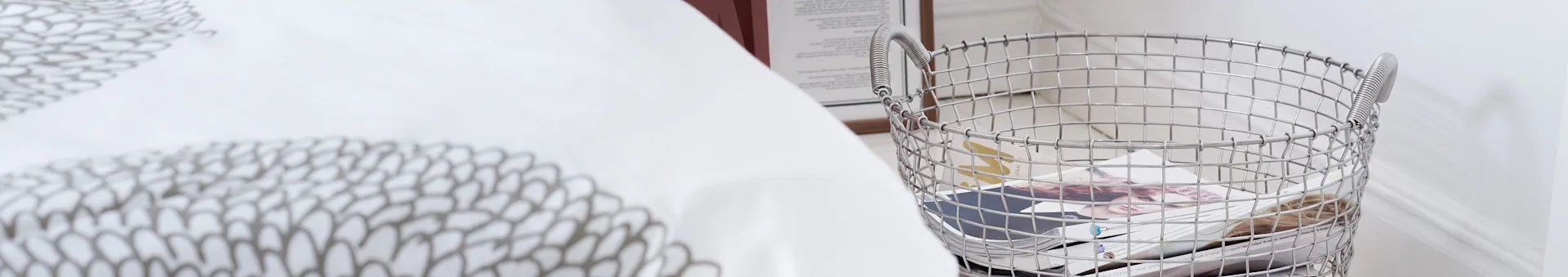 White patterned duvet and a silver wire basket holding magazines.