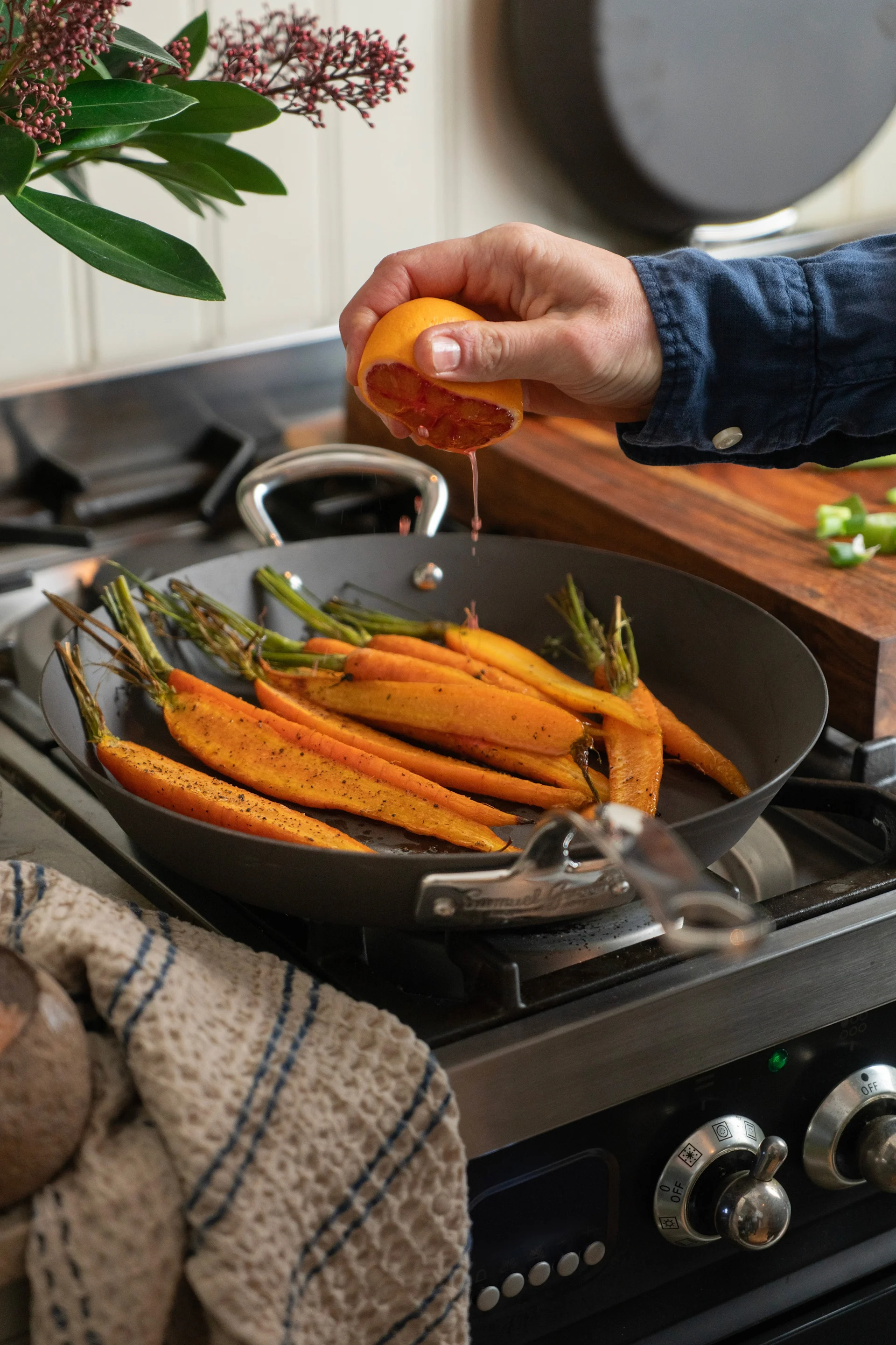 A hand squeezes a blood orange over a pan of roasted carrots on a gas stovetop.