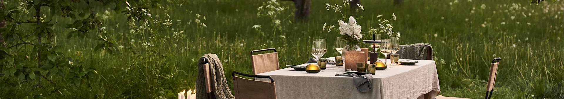 Outdoor dining table set with white flowers, glasses, and lamps in a lush green field.