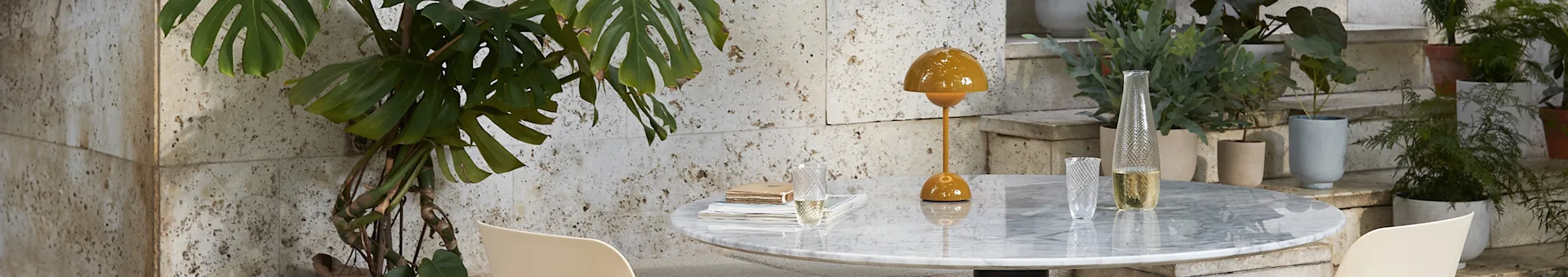 A mustard yellow table lamp and clear glass vase on a marble table with a monstera plant to the left and potted plants on stairs to the right.