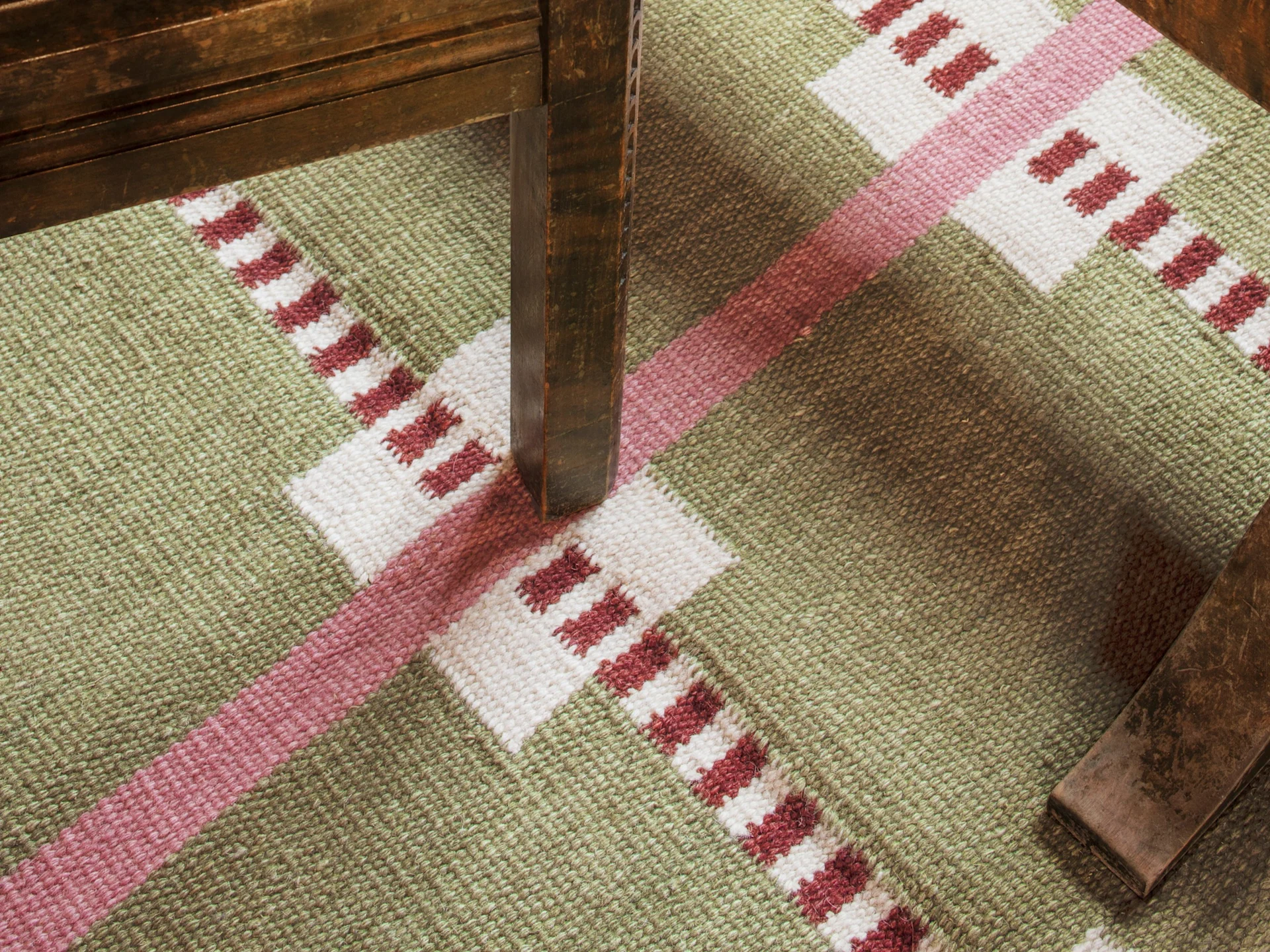 A green, white, and red geometric patterned rug with a dark wooden furniture leg casting a shadow.