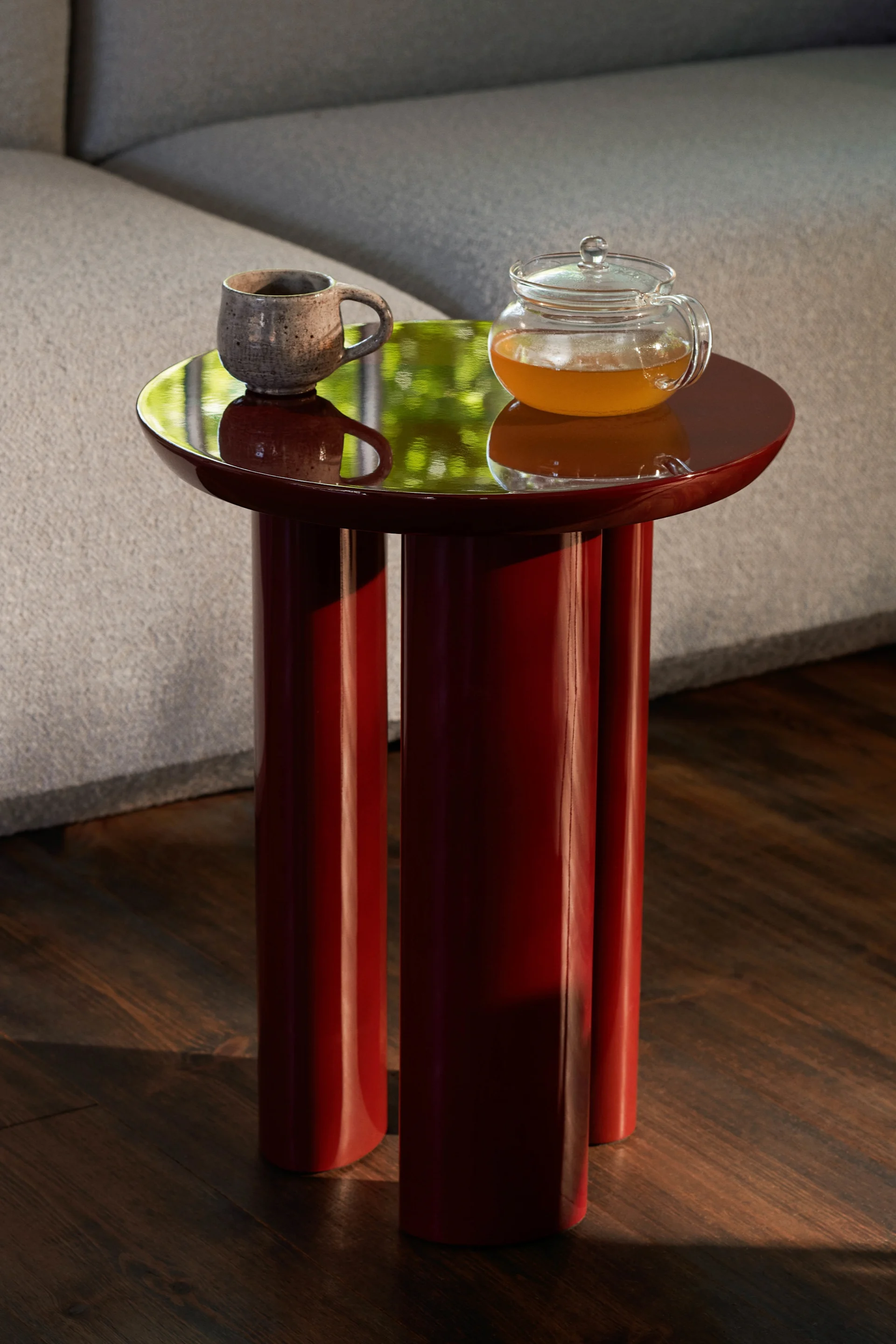 Dark red glossy side table with a speckled mug and glass teapot. Green reflections on top, beside a grey sofa and wood floor.