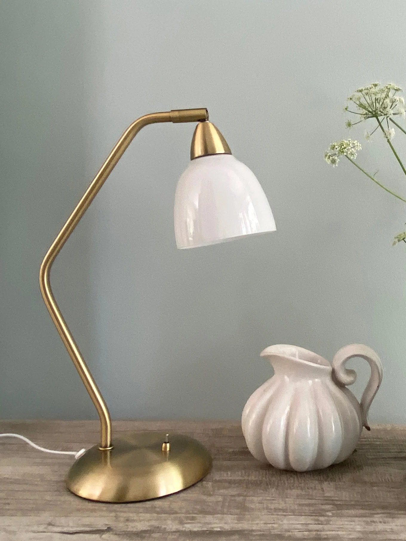 A brass table lamp with a white glass shade stands next to a white fluted ceramic pitcher on a wooden surface.